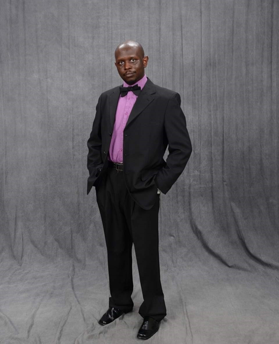 Man in a black suit with purple shirt and bow tie, standing against a gray backdrop.