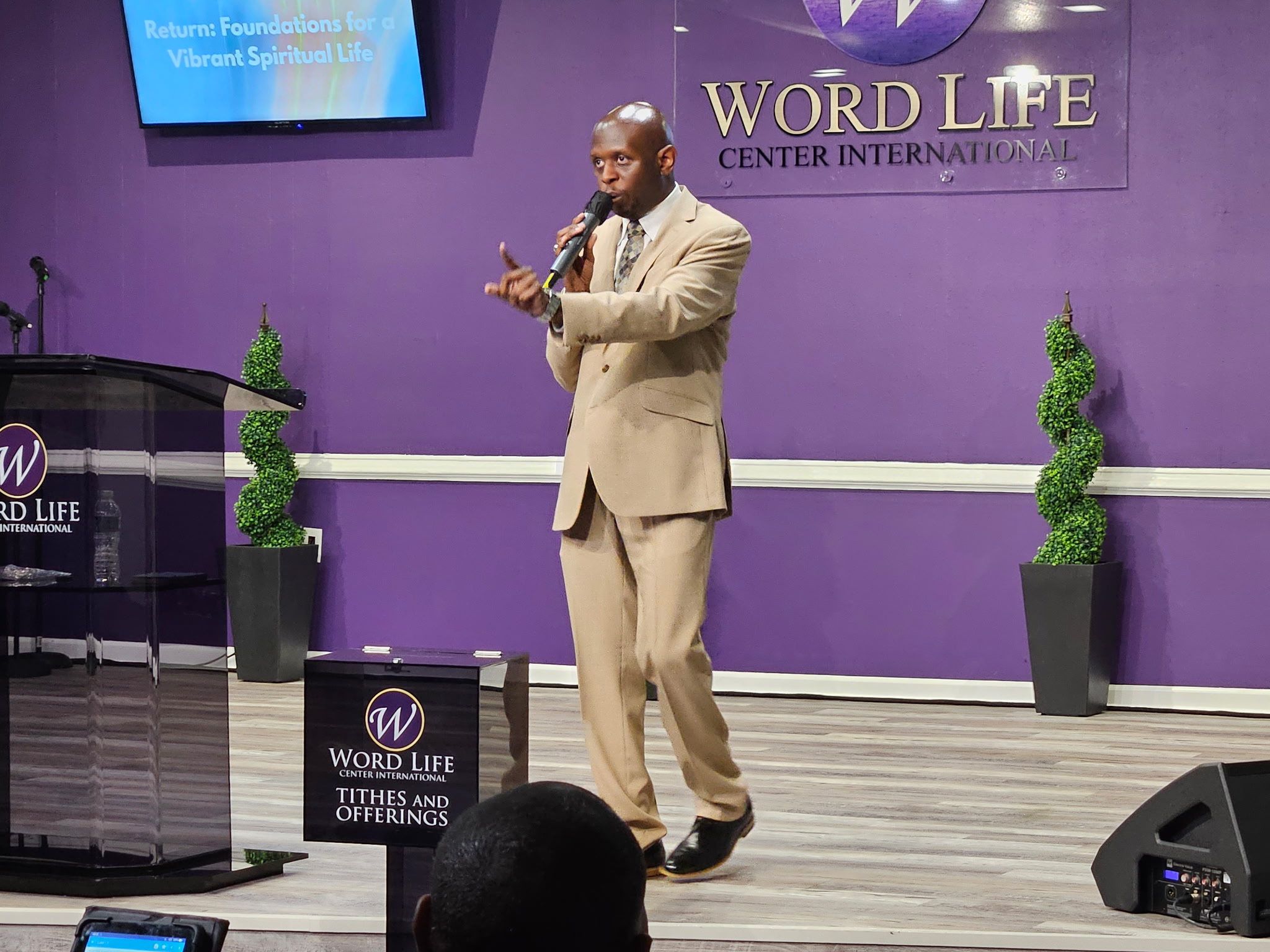 Man in beige suit speaking on stage with purple background and Word Life logo, two potted plants, and a podium.
