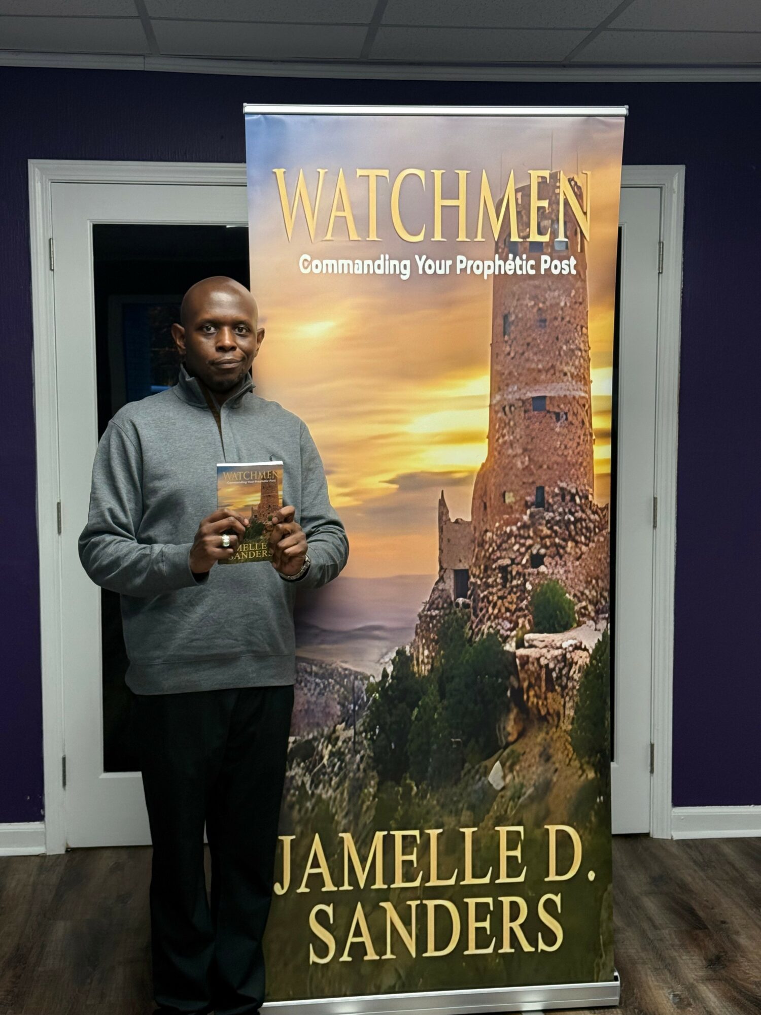 Man standing indoors holding a book in front of a large vertical banner with a castle and sunset background.