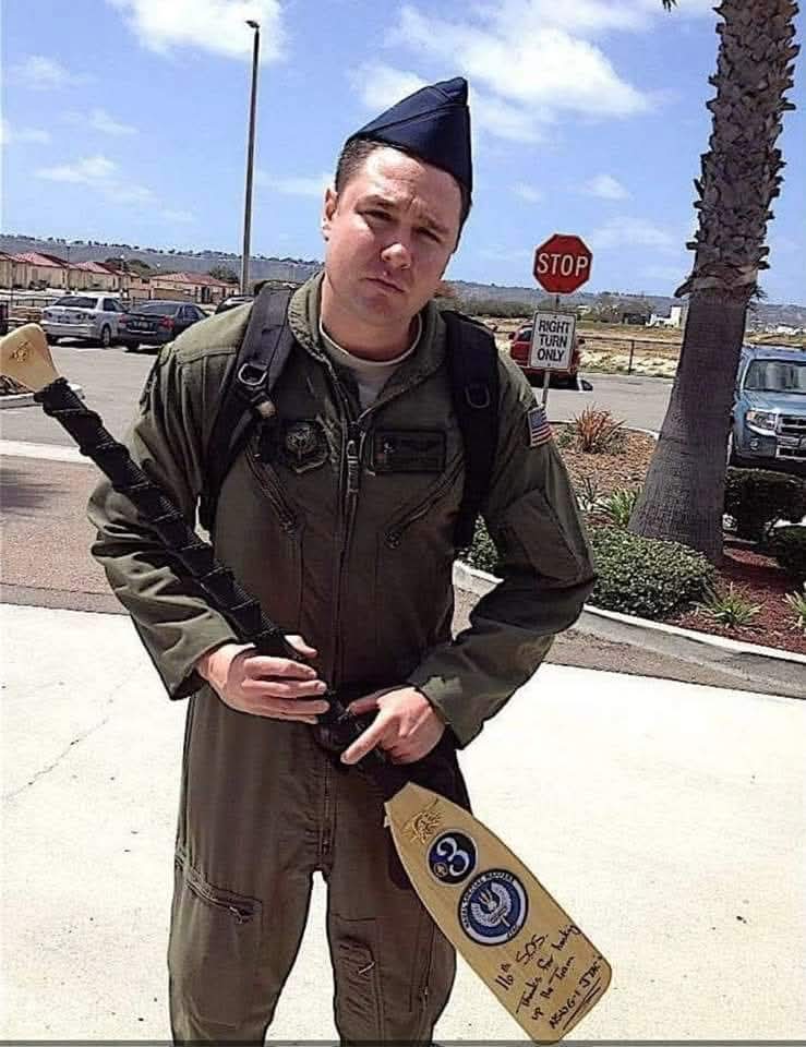 Person dressed in military uniform holding a rifle, standing outdoors near a stop sign and palm tree.