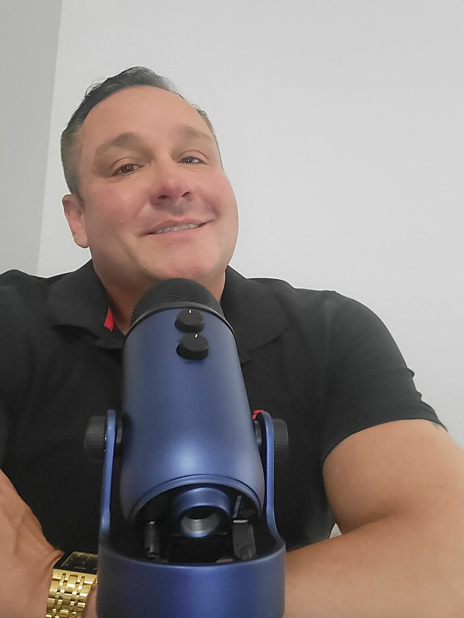 Man smiling at camera with a microphone in front, wearing a black shirt, against a plain white wall.
