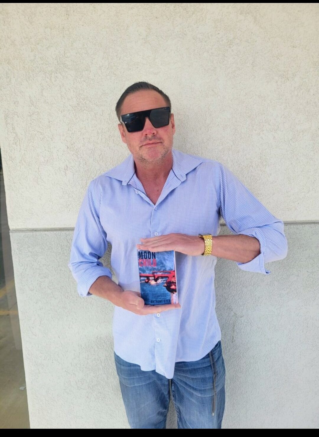 Man with sunglasses in light blue shirt holding a book, standing against a plain wall.