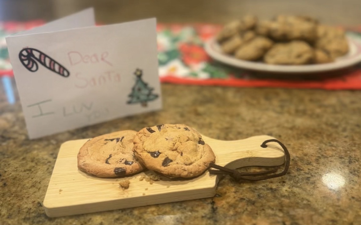 Two chocolate chip cookies on a small wooden tray with a metal handle, on a kitchen counter. A note and a plate of cookies are in the background.