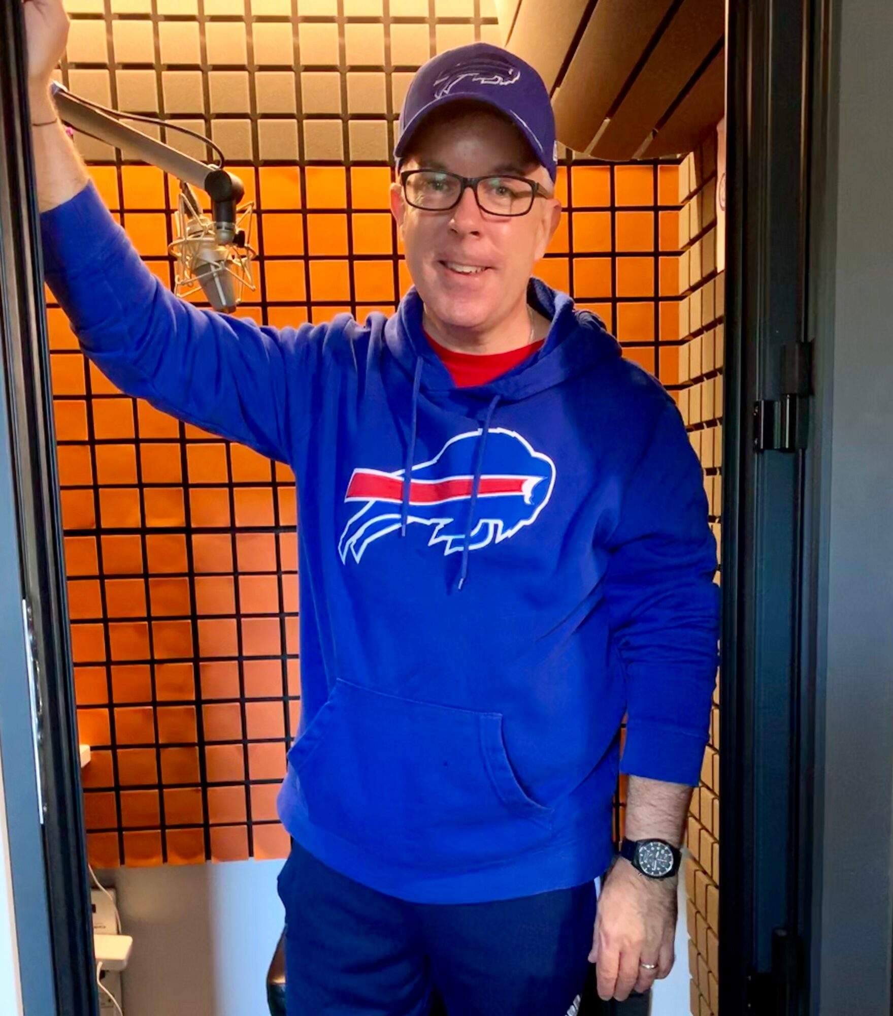 Person wearing glasses, a blue hoodie with a sports team logo, and a cap, standing in a booth with orange grid walls.