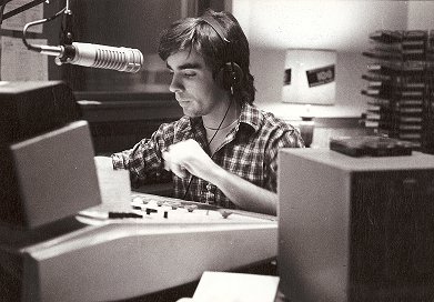 Person with headphones working at a radio station with equipment and stacks of papers nearby.