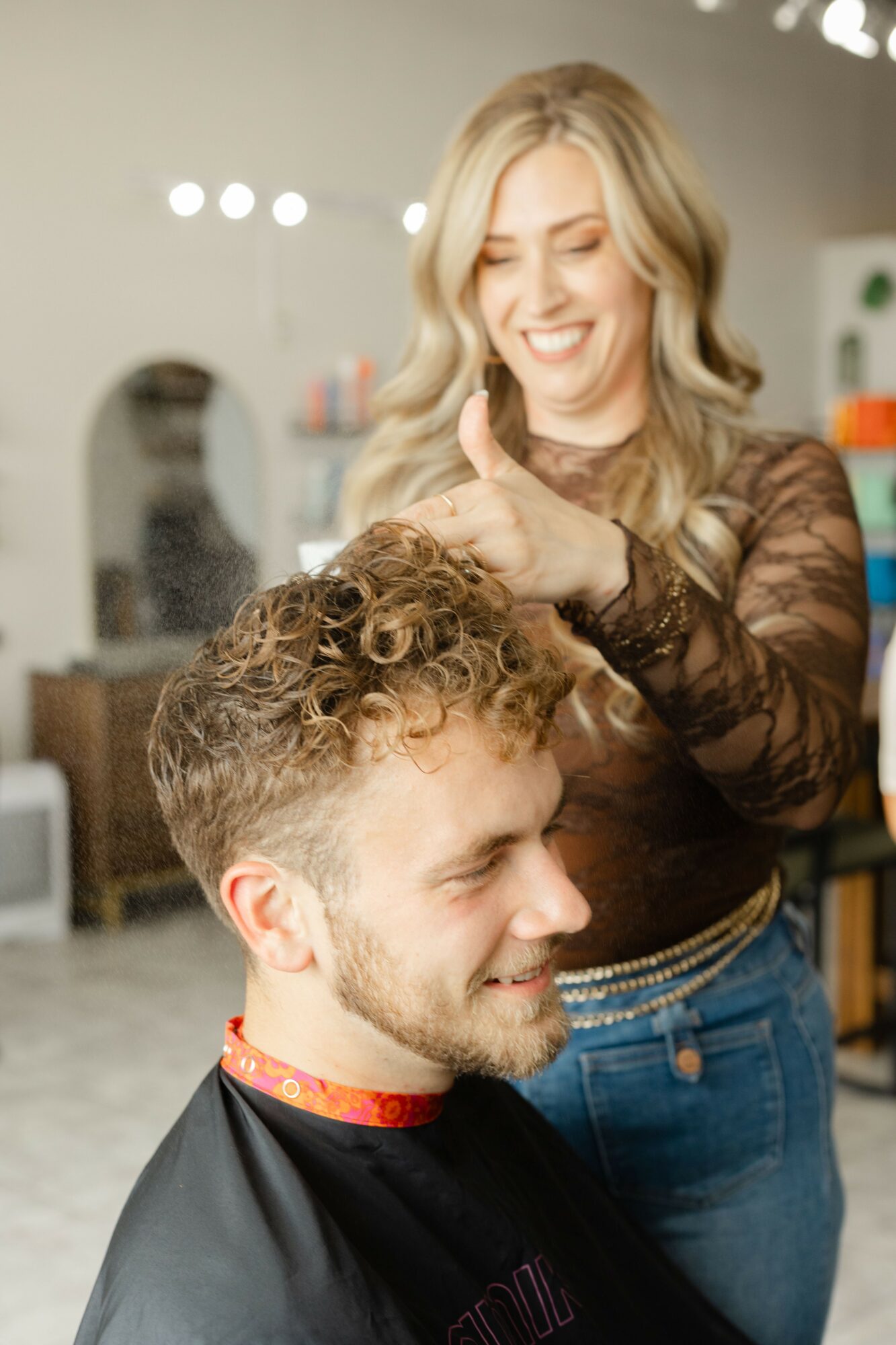 Woman with blonde hair smiling and giving a thumbs-up behind a man with curly hair in a salon.