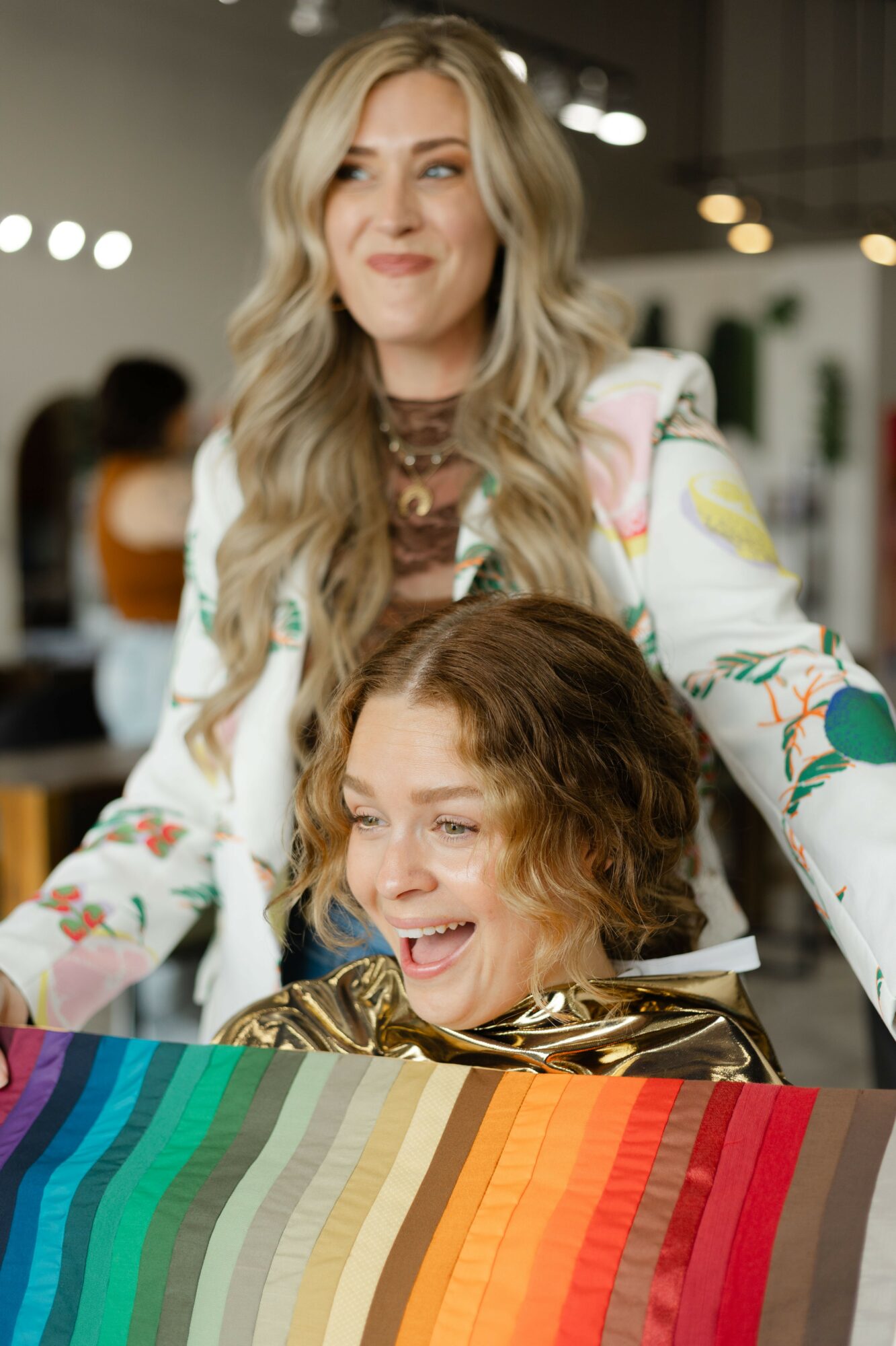 Two women smiling, one with long blonde hair and the other with curly brown hair, in an indoor setting.