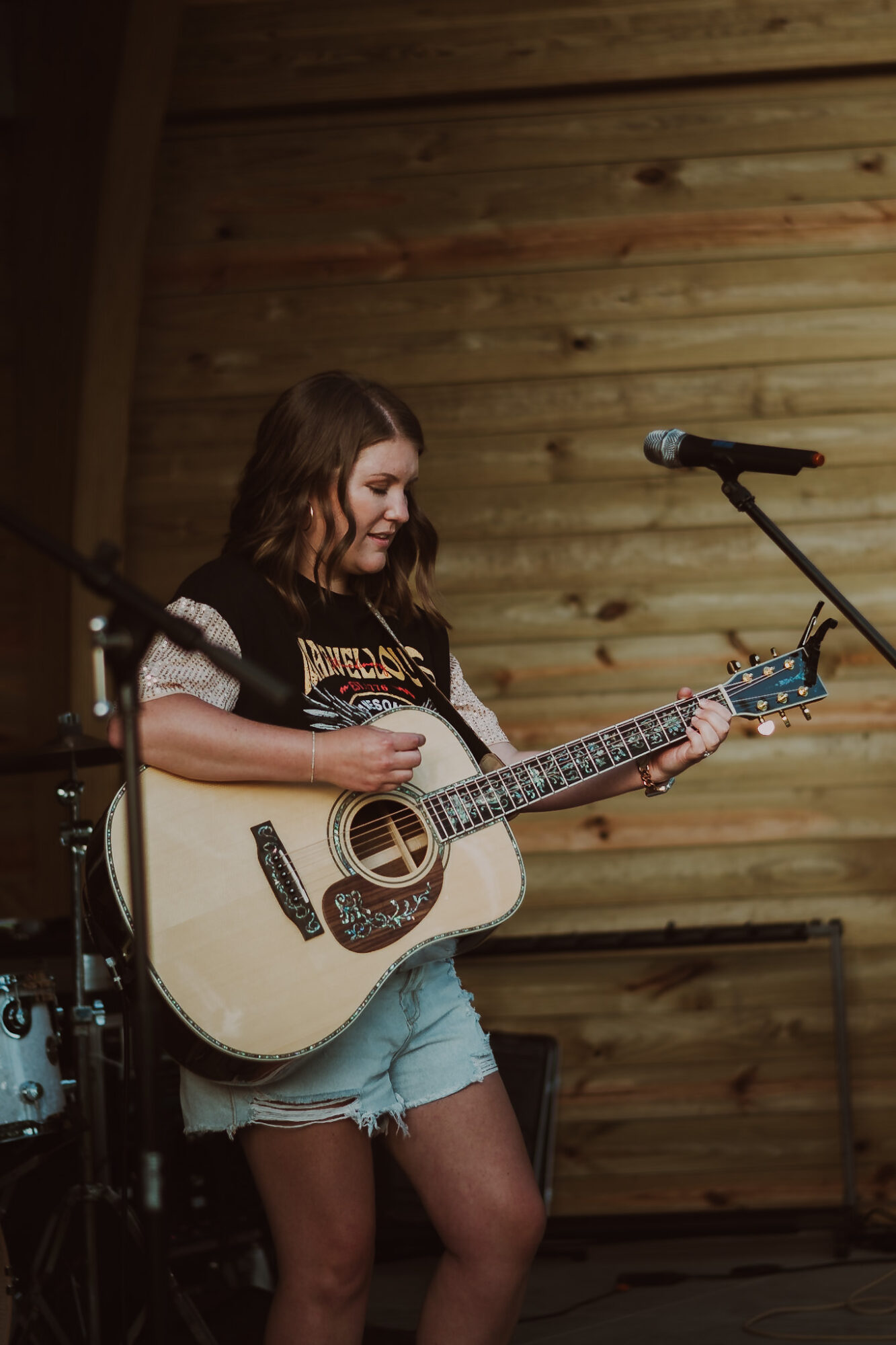 Young woman playing an acoustic guitar on stage with wooden background and microphones nearby.