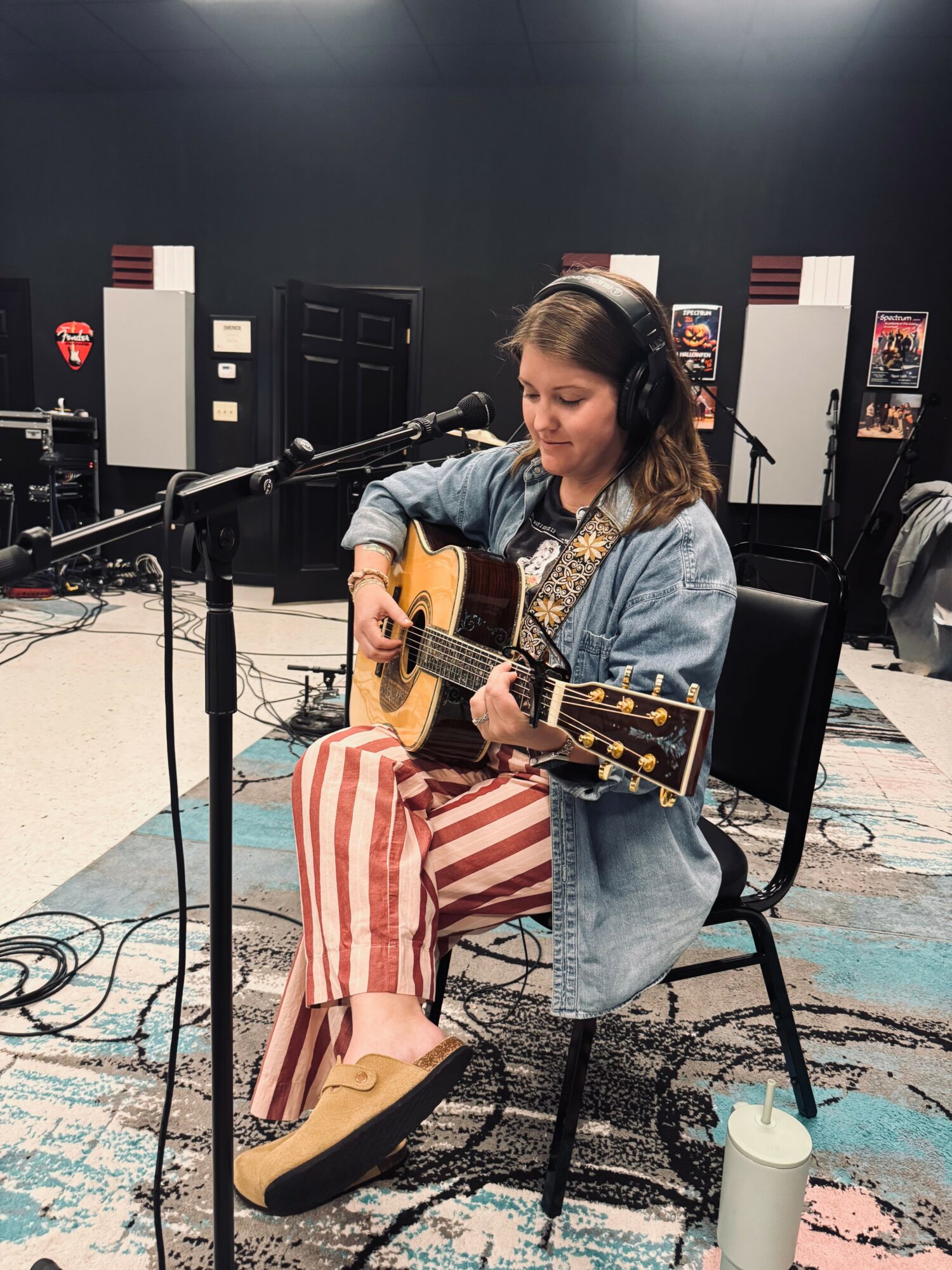 Young woman with headphones playing acoustic guitar in a recording studio, seated on a black chair, with equipment and posters in background.