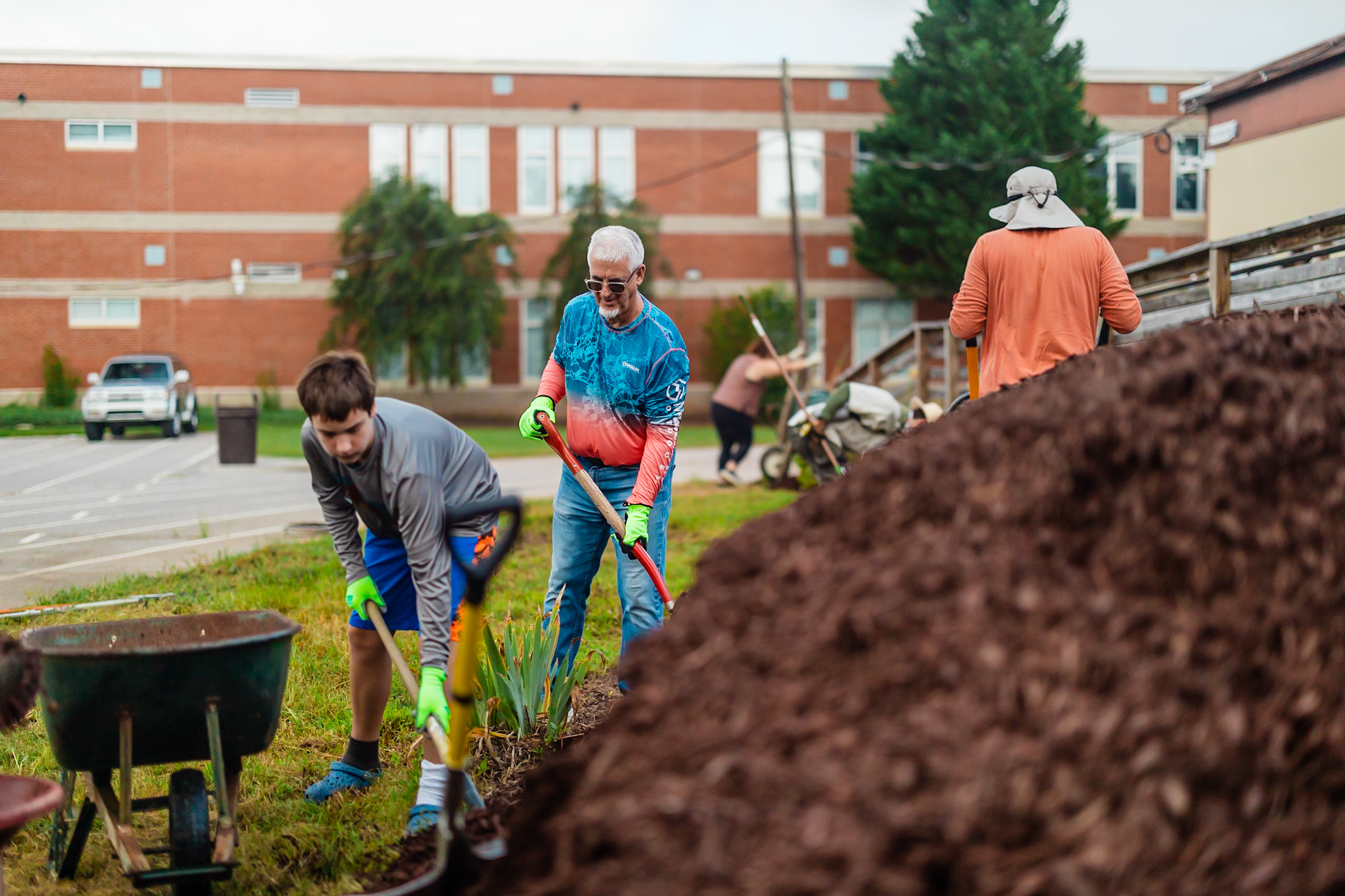 People planting flowers outdoors with a large mound of soil in foreground, building and trees in background.