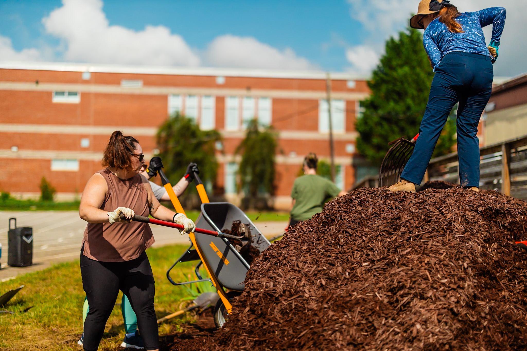 Two women working outdoors near a large pile of mulch, with a brick building and trees in the background.