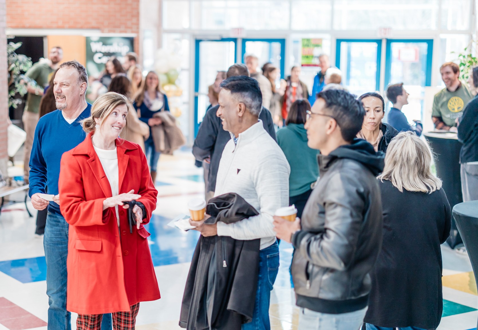 Group of people socializing indoors, some holding drinks, in a bright, modern space with large windows.