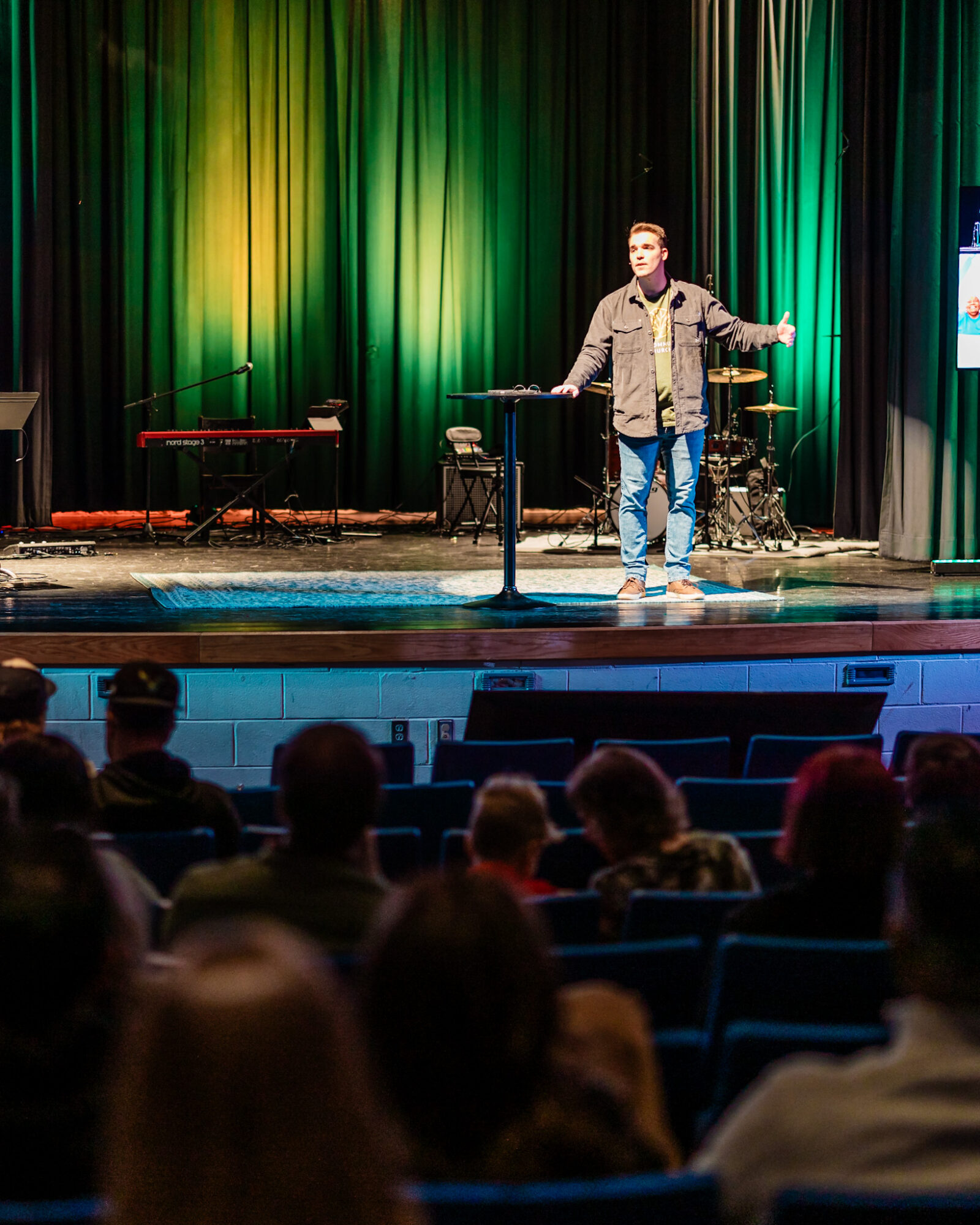 Person standing on stage with arms outstretched, colorful curtains in background, audience seated watching.