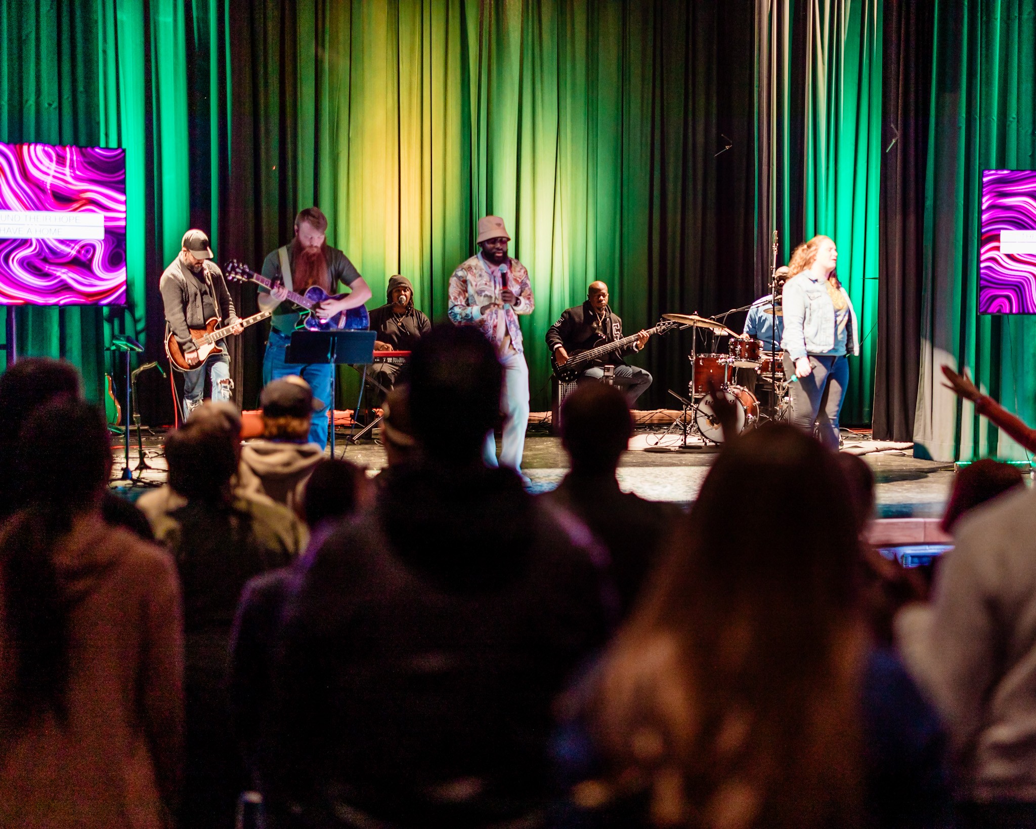 Band performs on stage with colorful curtains, audience watching attentively, some with heads turned towards the stage.