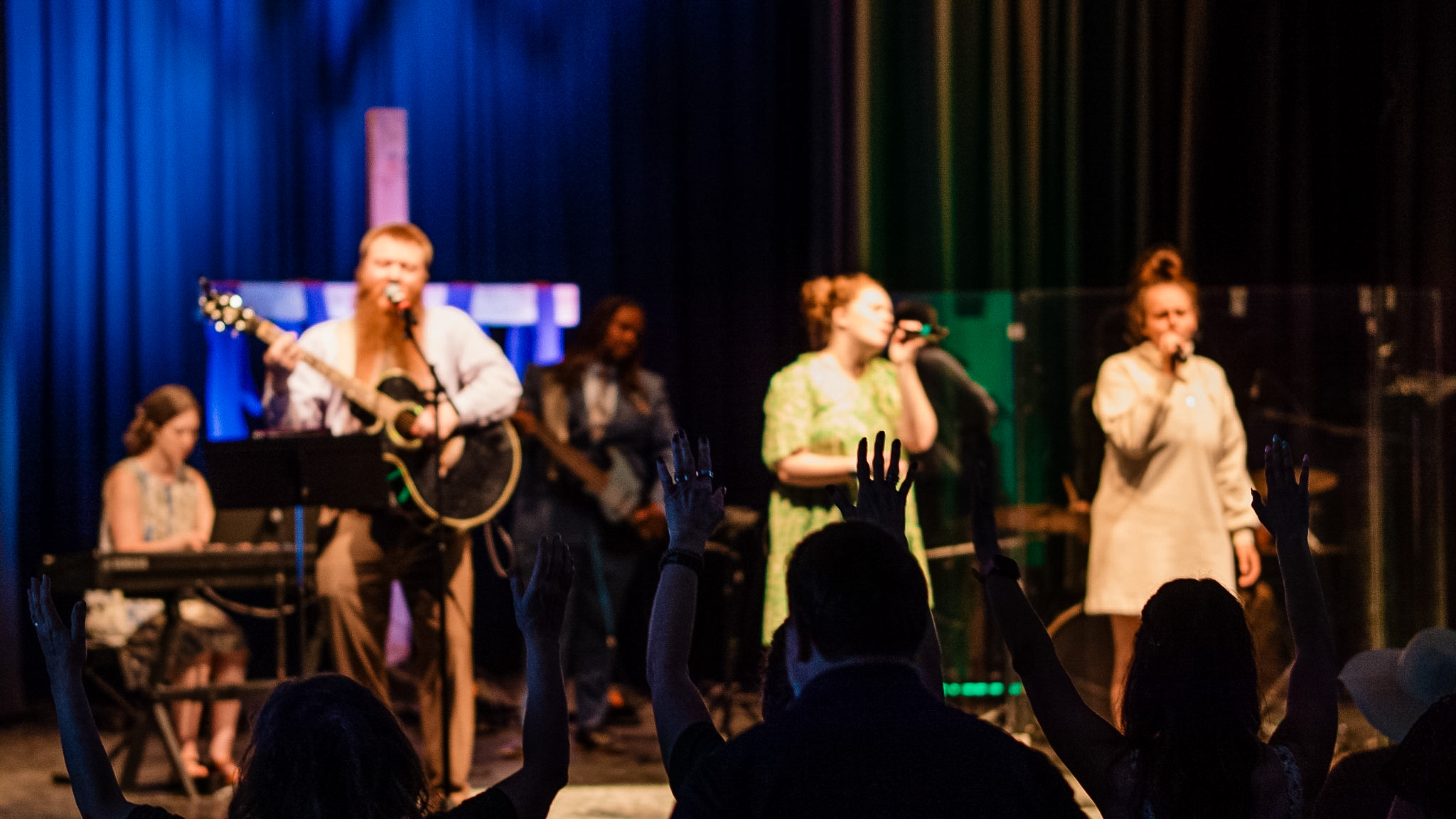 Group of people performing on stage with microphones and musical instruments, audience visible in foreground.