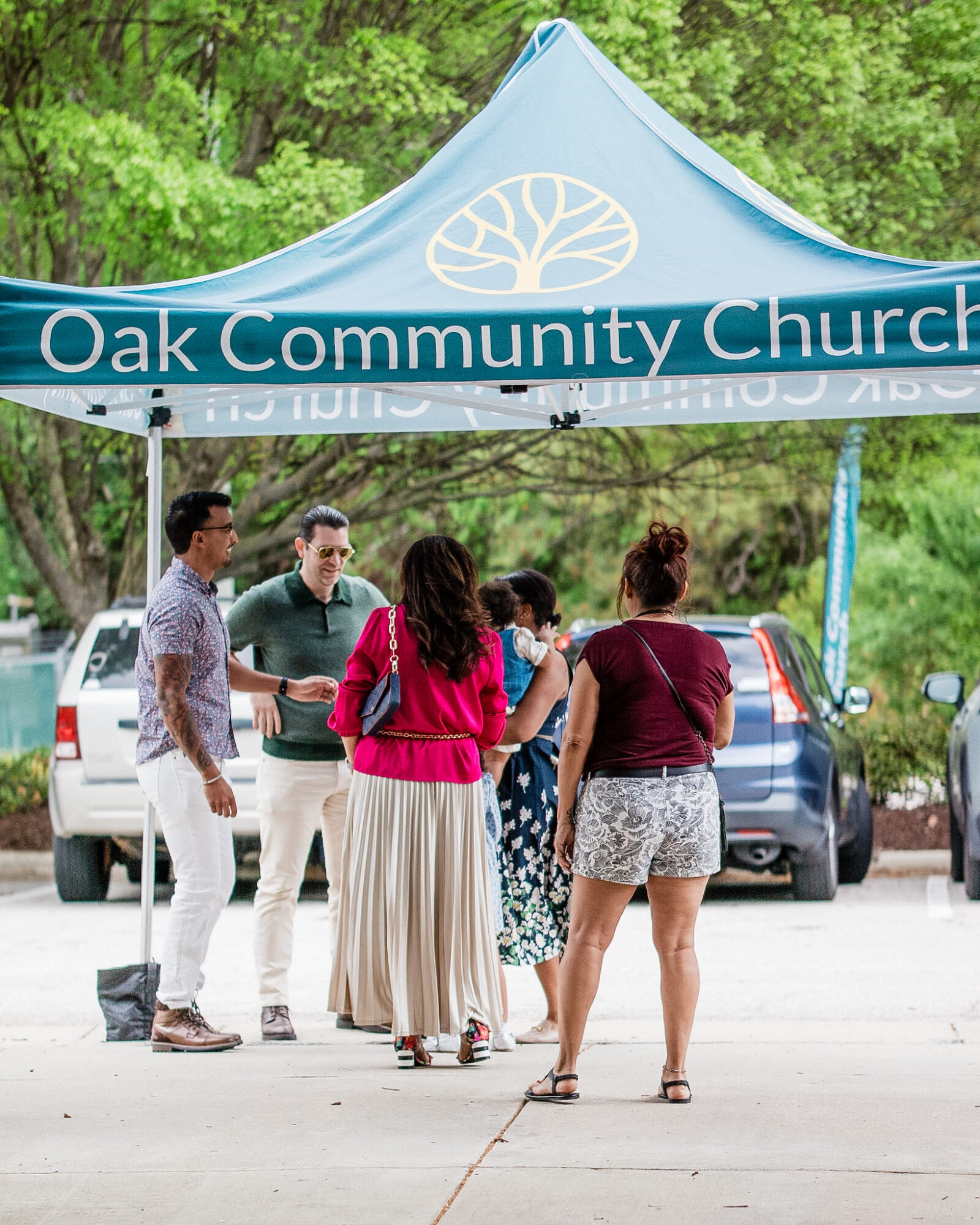 Group of people standing under a blue tent with trees in background, near parked cars.