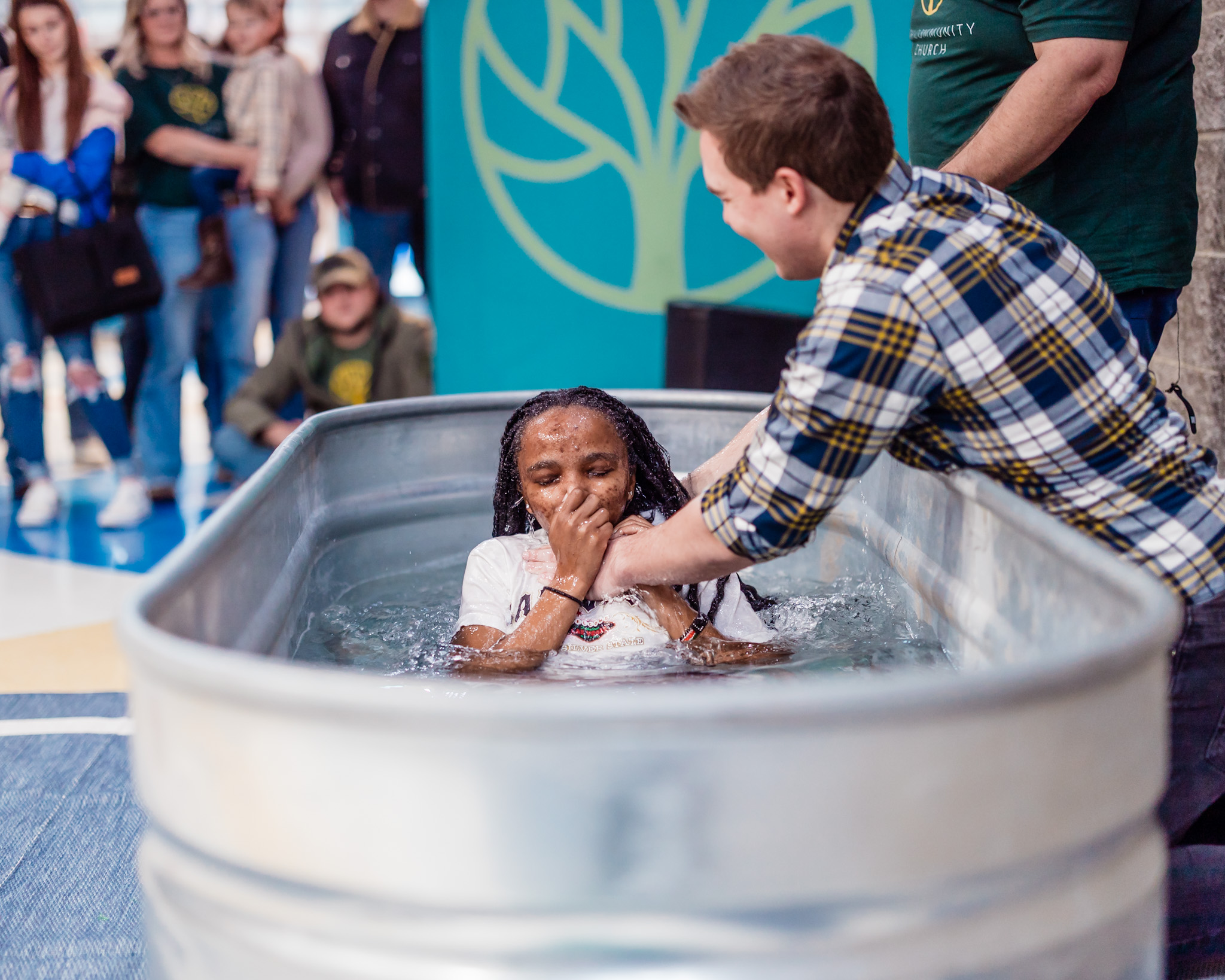 Young girl in a metal tub, water, covering nose with hand, person pouring water over her head, group of people in background.