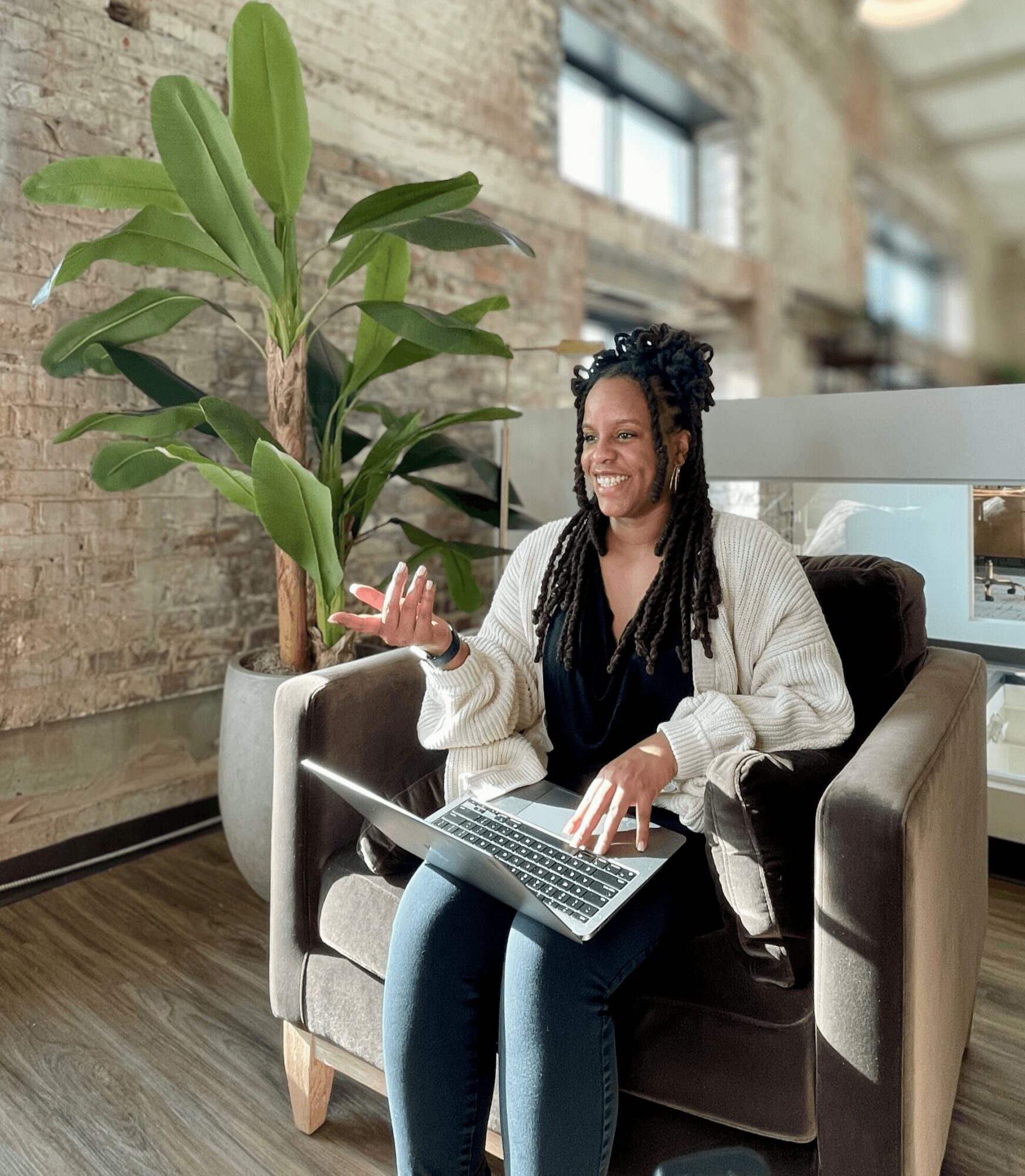 Woman sitting on armchair with laptop, smiling, in industrial-style room with large window and green plant.