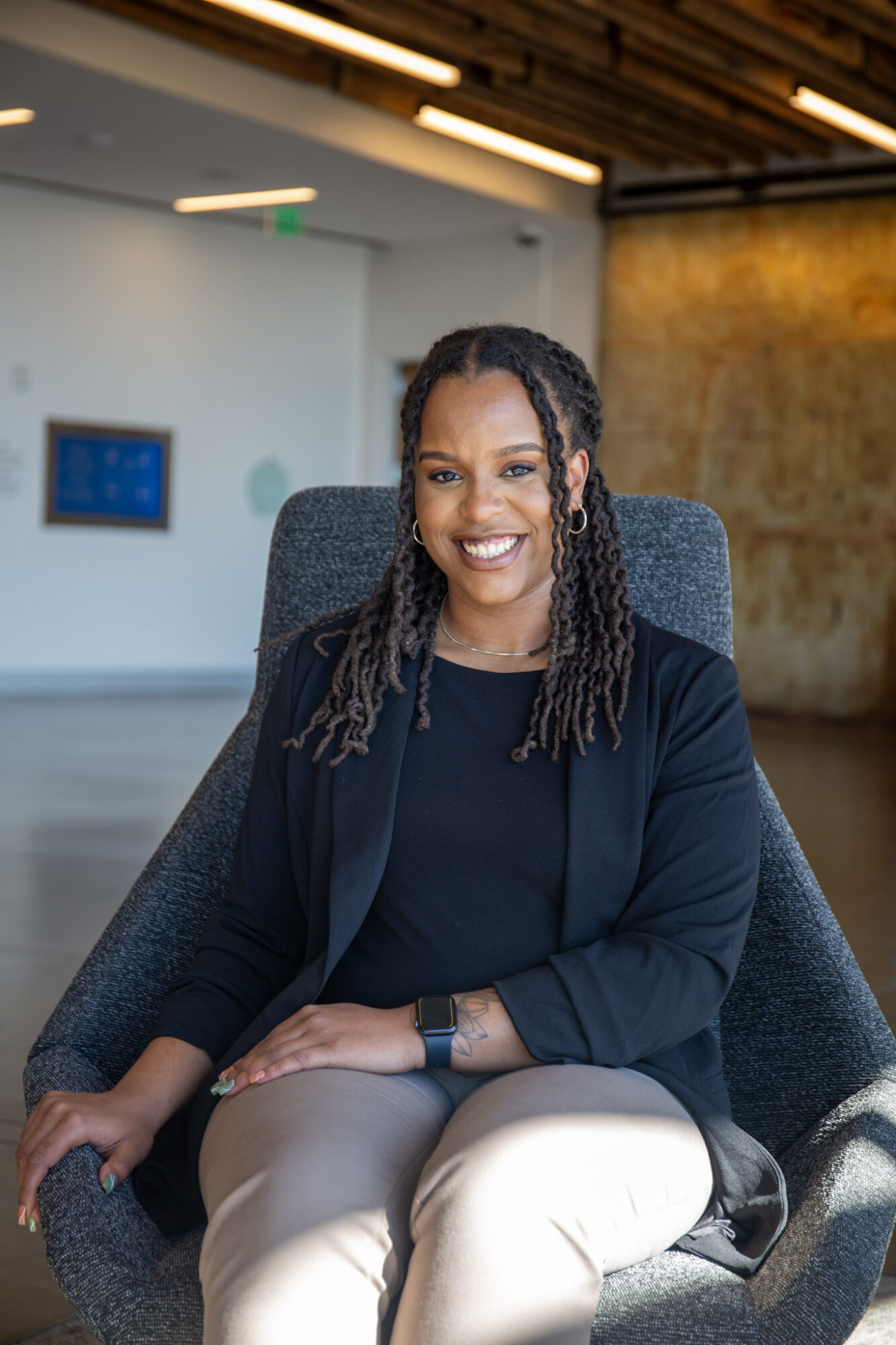 Smiling woman with dreadlocks sitting in a modern office chair, wearing a dark blazer and light-colored pants.