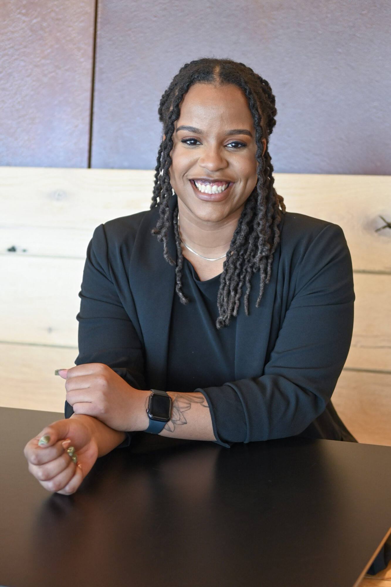 Smiling woman with braided hair, wearing a black top, sitting at a table in a modern setting.