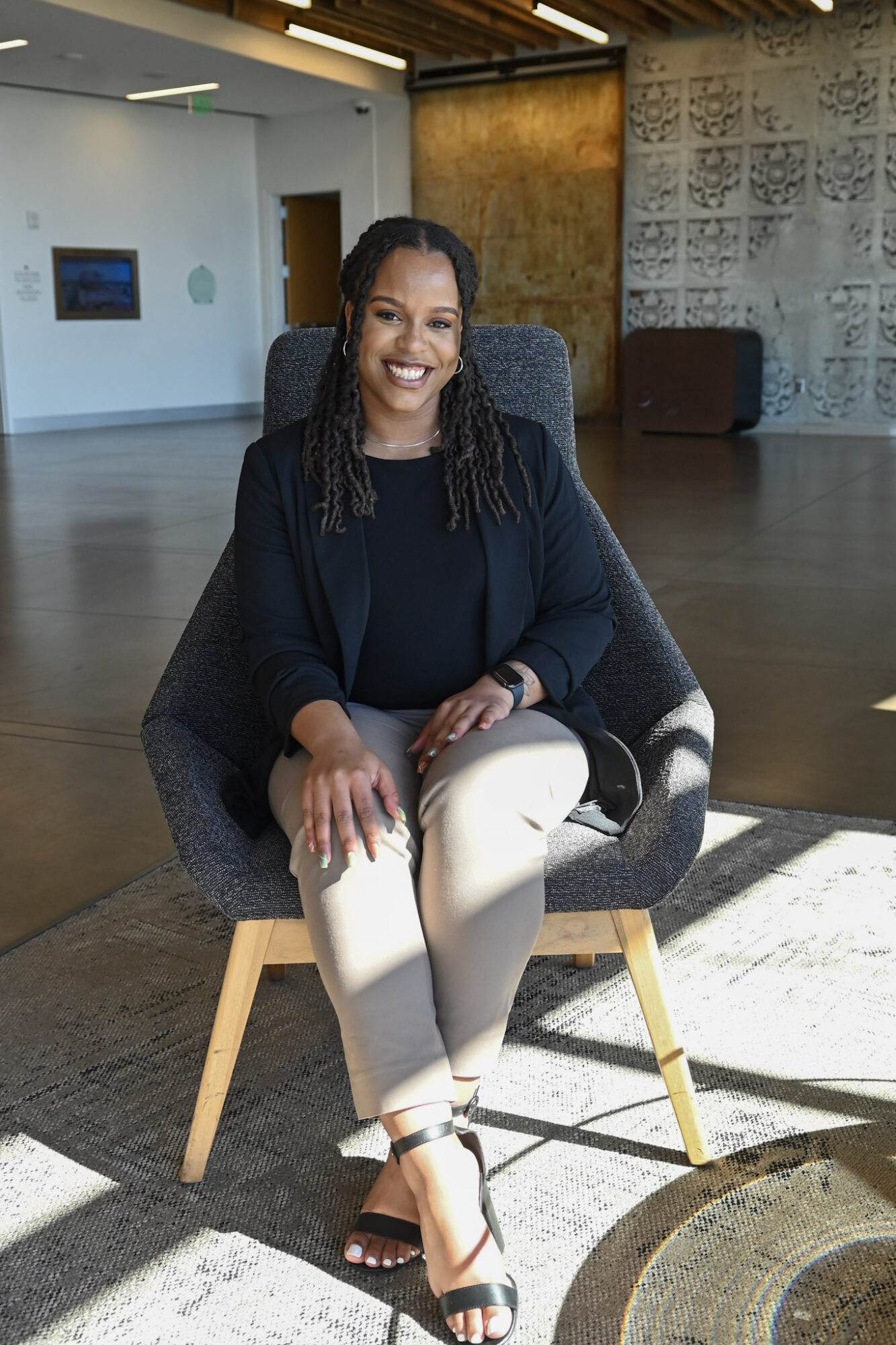 Woman sitting in a chair, smiling, in a modern indoor space with sunlight and decorative wall.