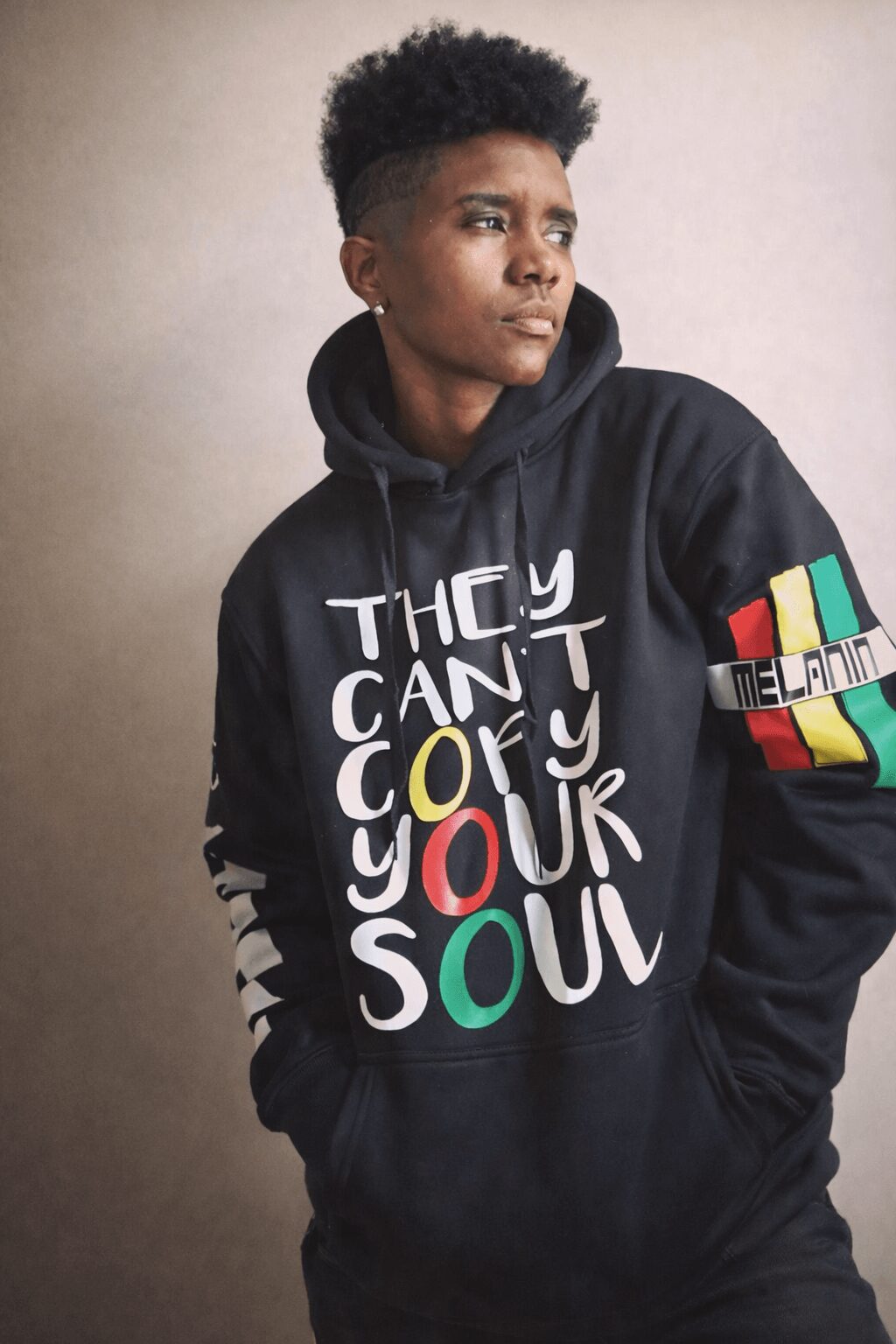 Young person with short curly hair wearing a black hoodie with colorful text and design, standing against a plain background.