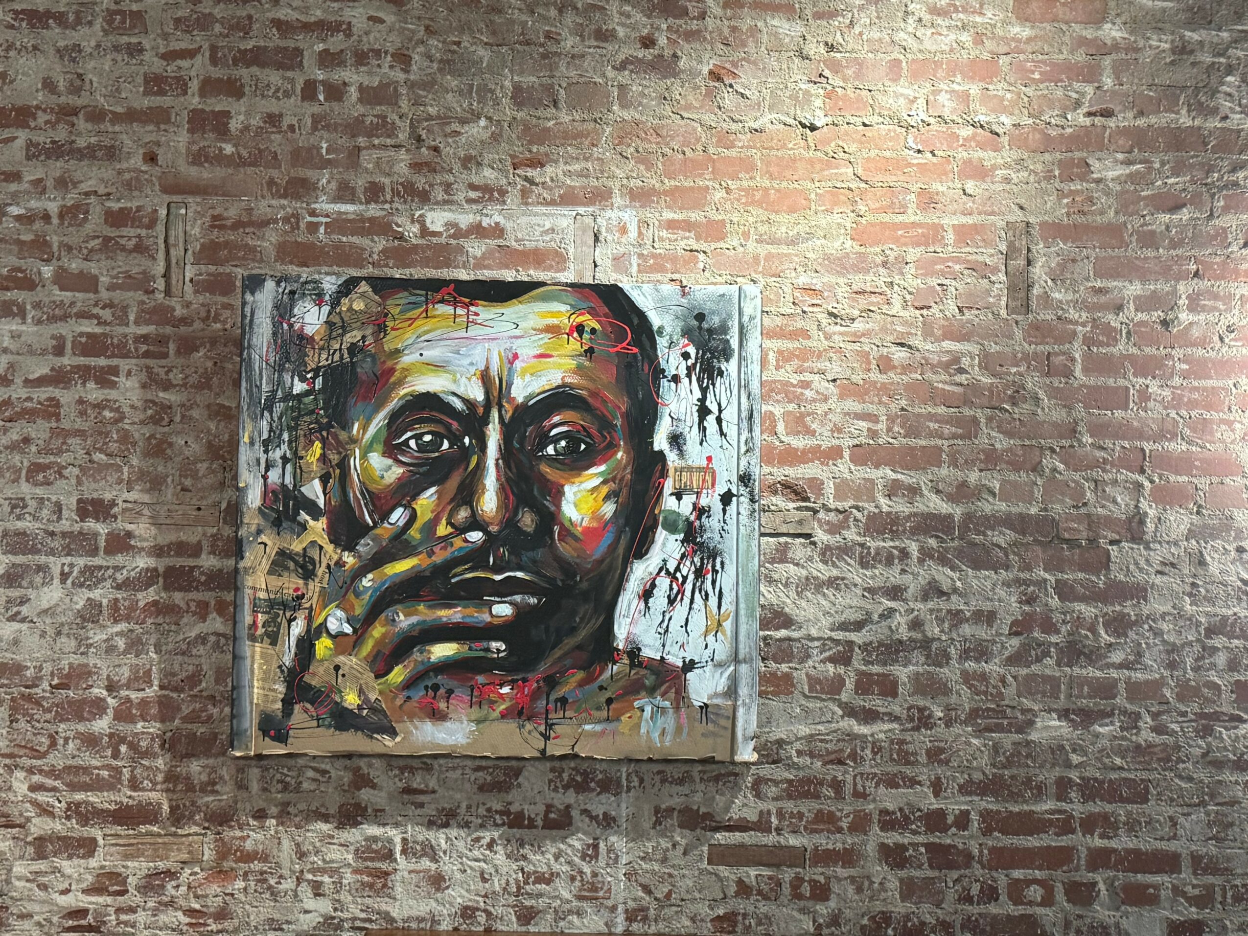 Colorful portrait of a person with hand near face, painted on canvas against brick wall.