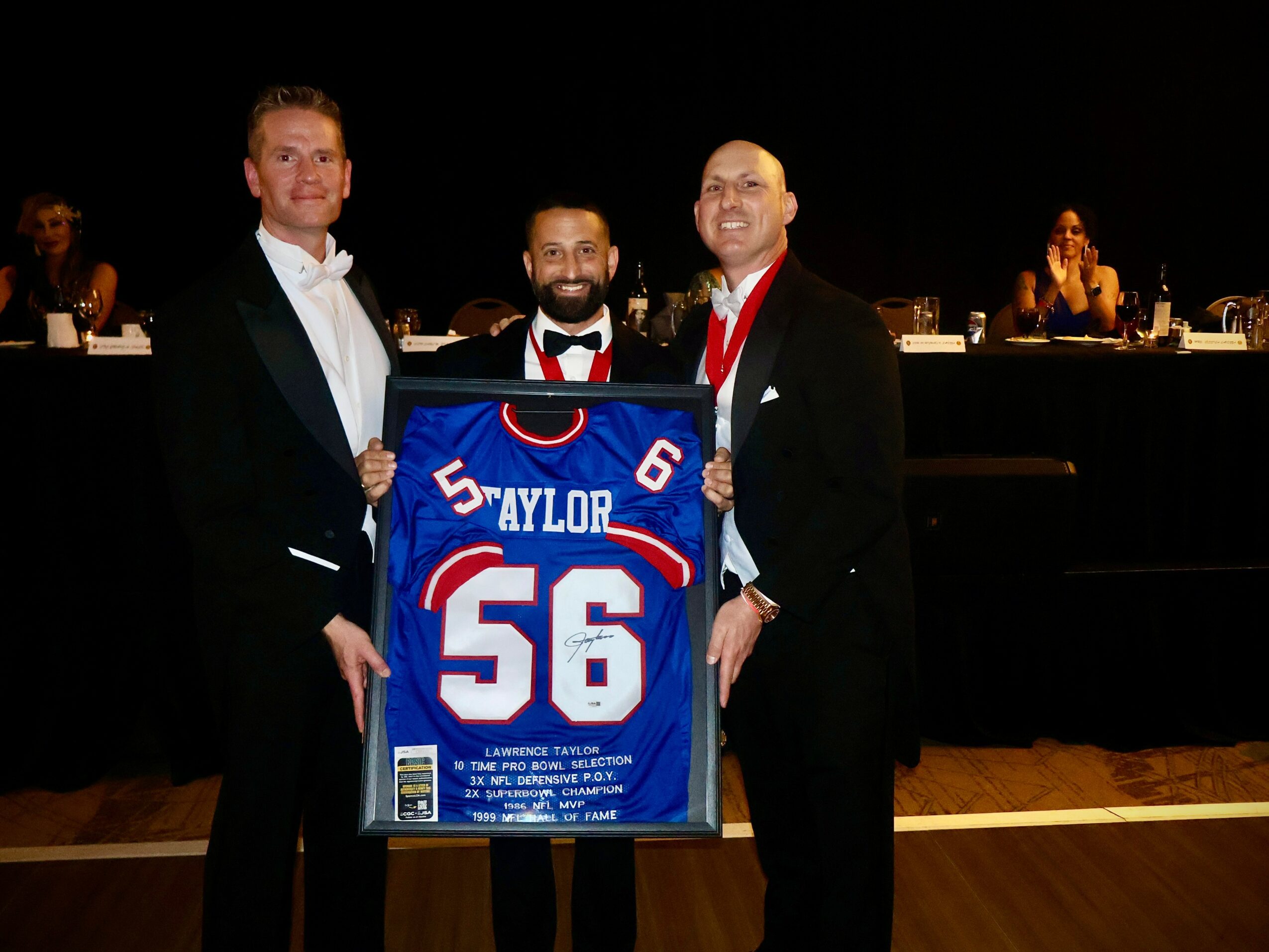 Three men in formal attire holding a framed sports jersey, with a dark background and seated people behind them.