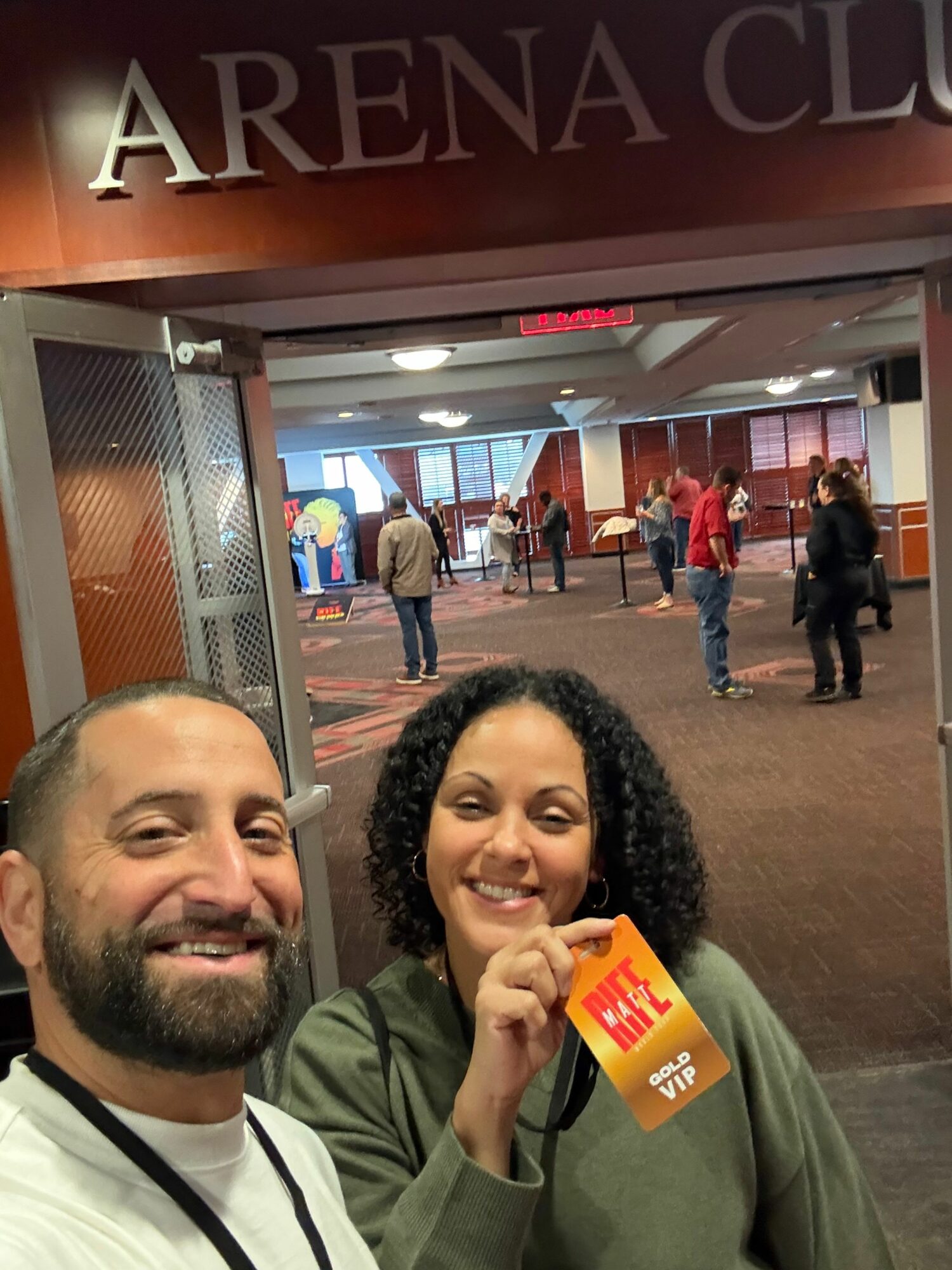 Two people smiling inside a building, with a lobby and people walking in the background.