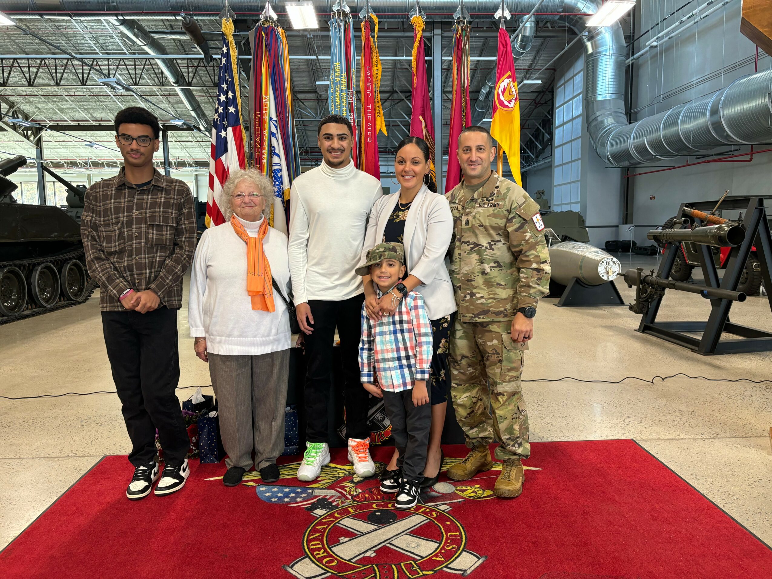 Group of six people, including a child, standing on a red carpet with flags behind them inside a building.