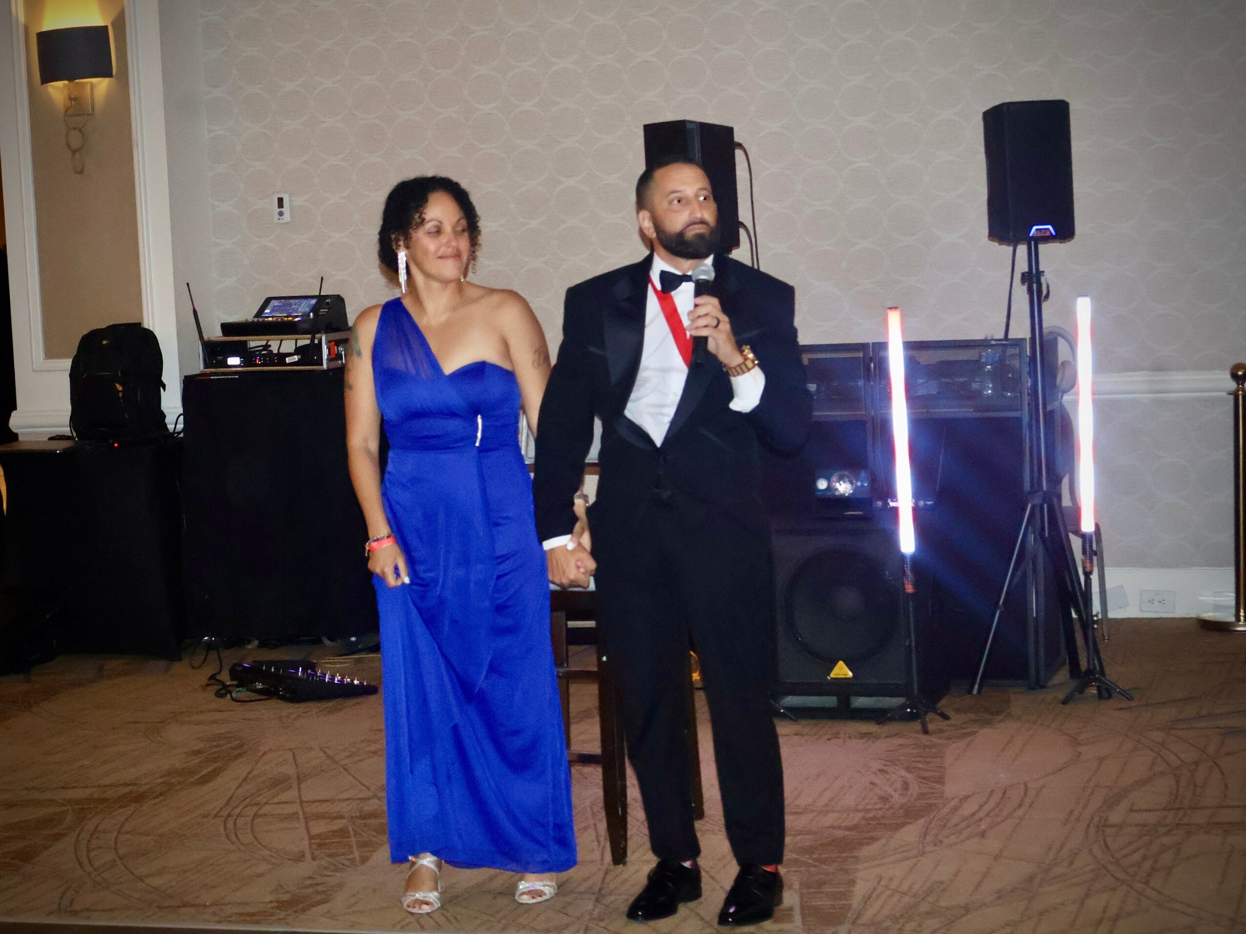 Man and woman holding hands, man speaking into microphone, in formal attire, in event hall with sound equipment.