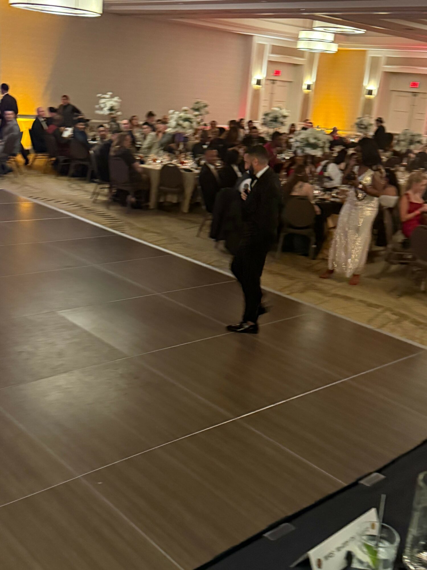 Person in formal attire standing on a dance floor at a banquet hall with round tables and guests in the background.