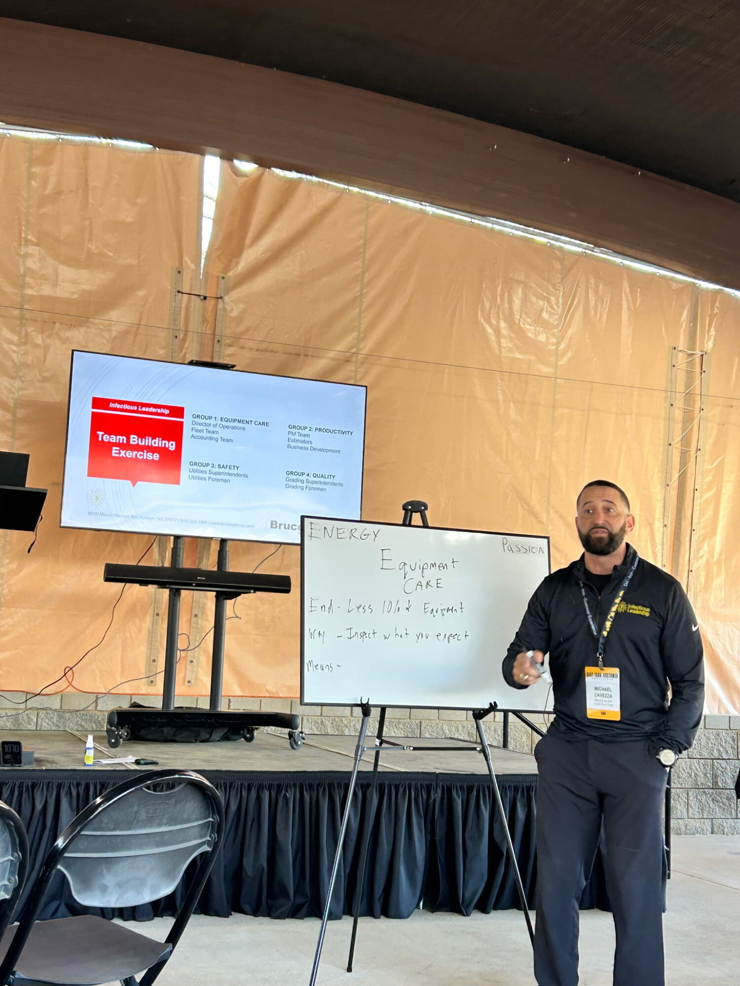 Man standing next to a whiteboard and a screen on stage at an indoor event.