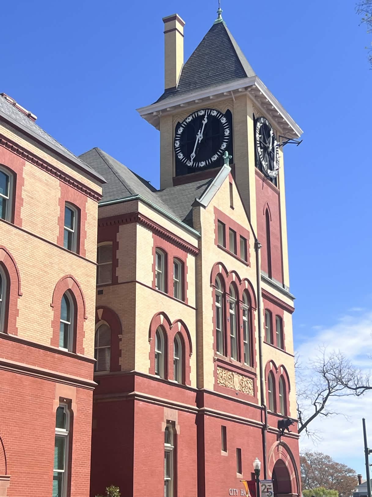Clock tower with a large clock face, part of a historic building, against a blue sky with a tree nearby.