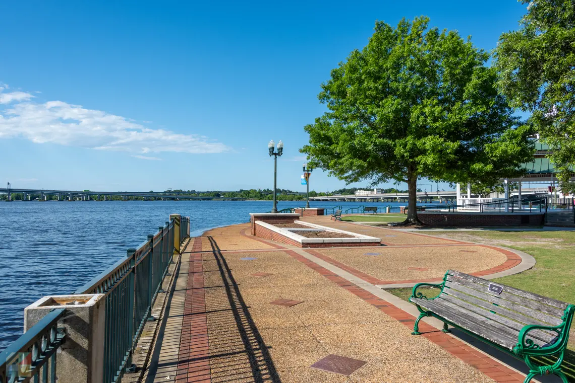 Waterfront promenade with trees, bench, and railing, overlooking a body of water under a blue sky.