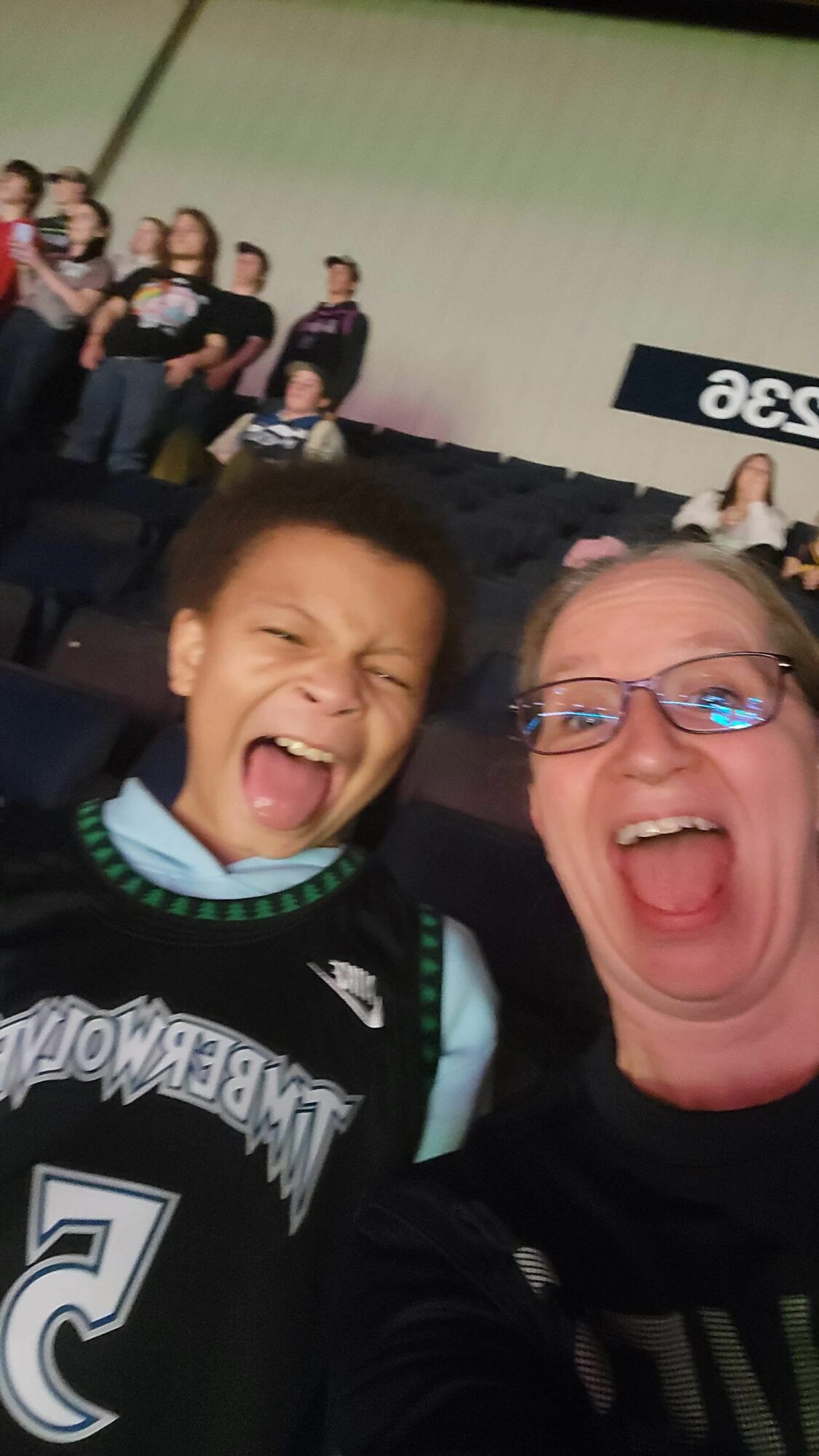 Two people smiling and taking a selfie at an indoor event with a group of people in the background.