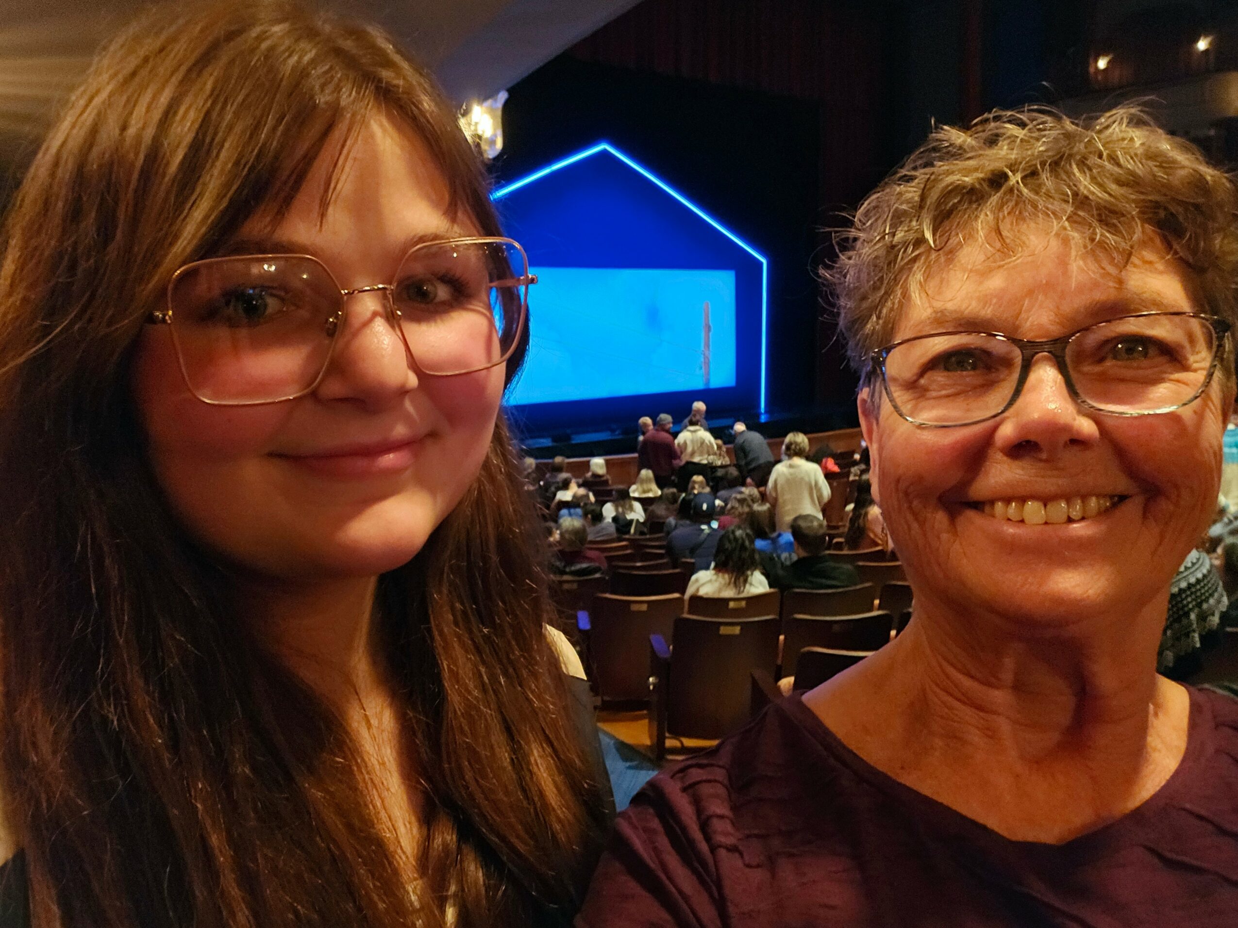 Two women smiling in a theater with an audience and a blue-lit stage in the background.