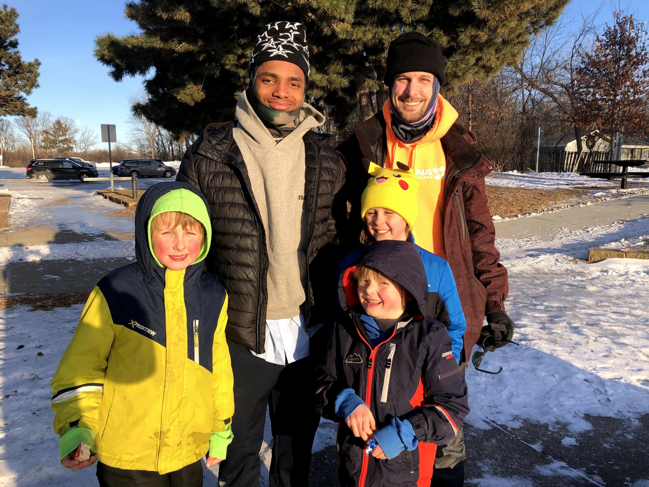 Two adults and three children outdoors in winter, standing on snow with trees and parked cars in background.