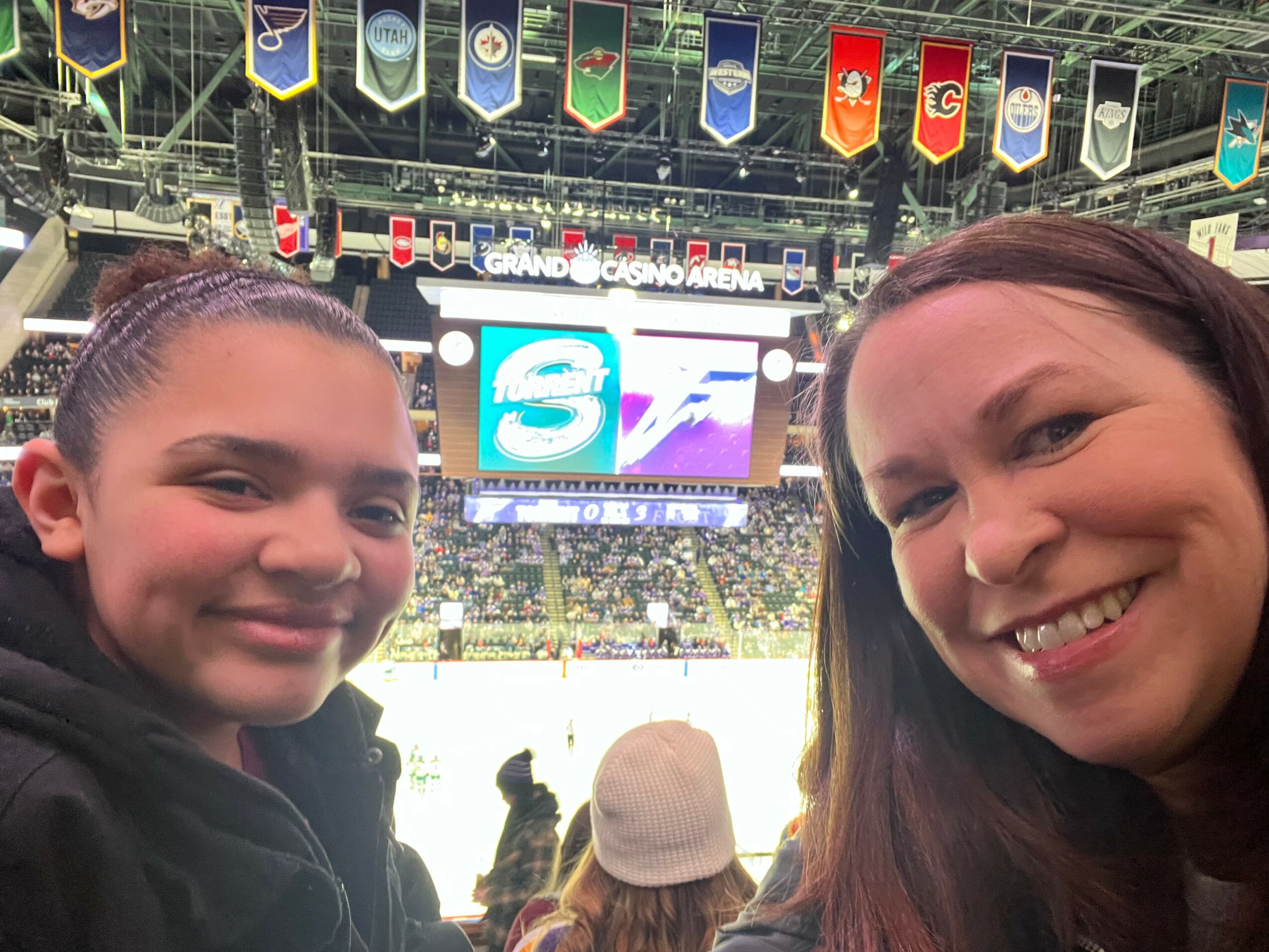 Two smiling women taking a selfie inside a sports arena with banners hanging from the ceiling and a large screen in the background.