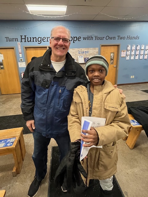 Two people standing indoors, smiling, with a blue wall and wooden benches in the background.