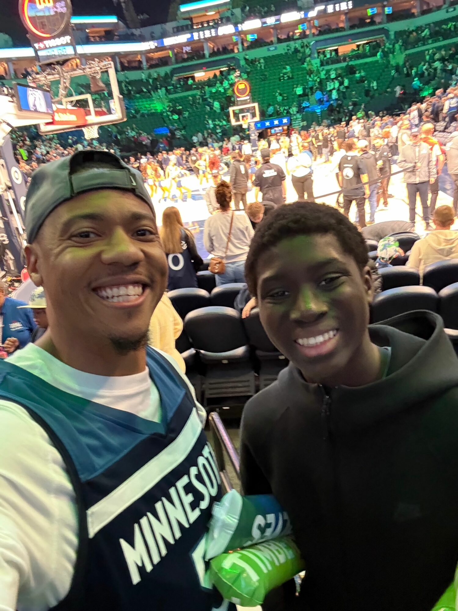Two smiling people taking a selfie at a basketball game with a crowd and court in the background.