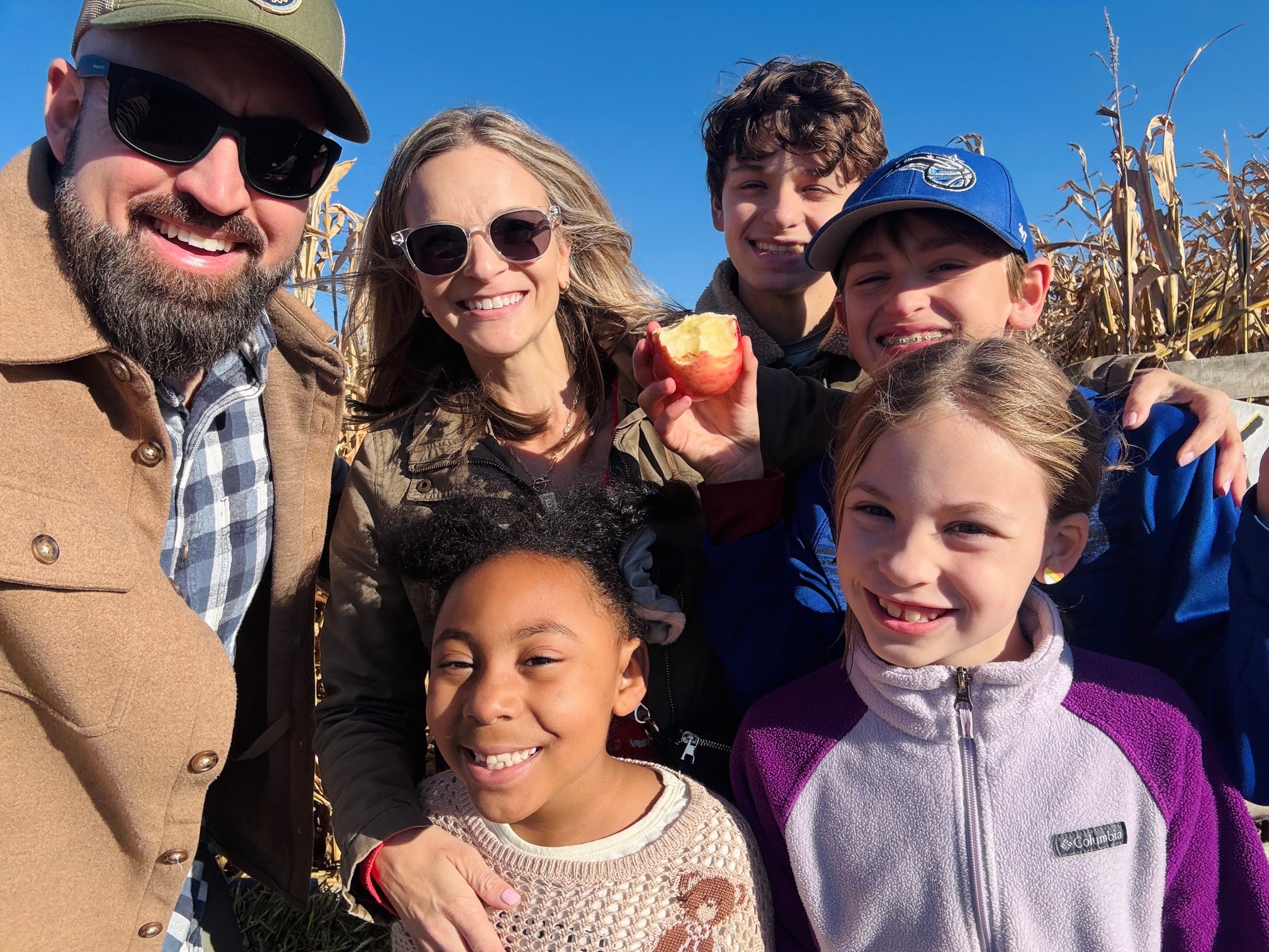 Group of six people smiling outdoors, holding apples, with blue sky and dried plants in background.