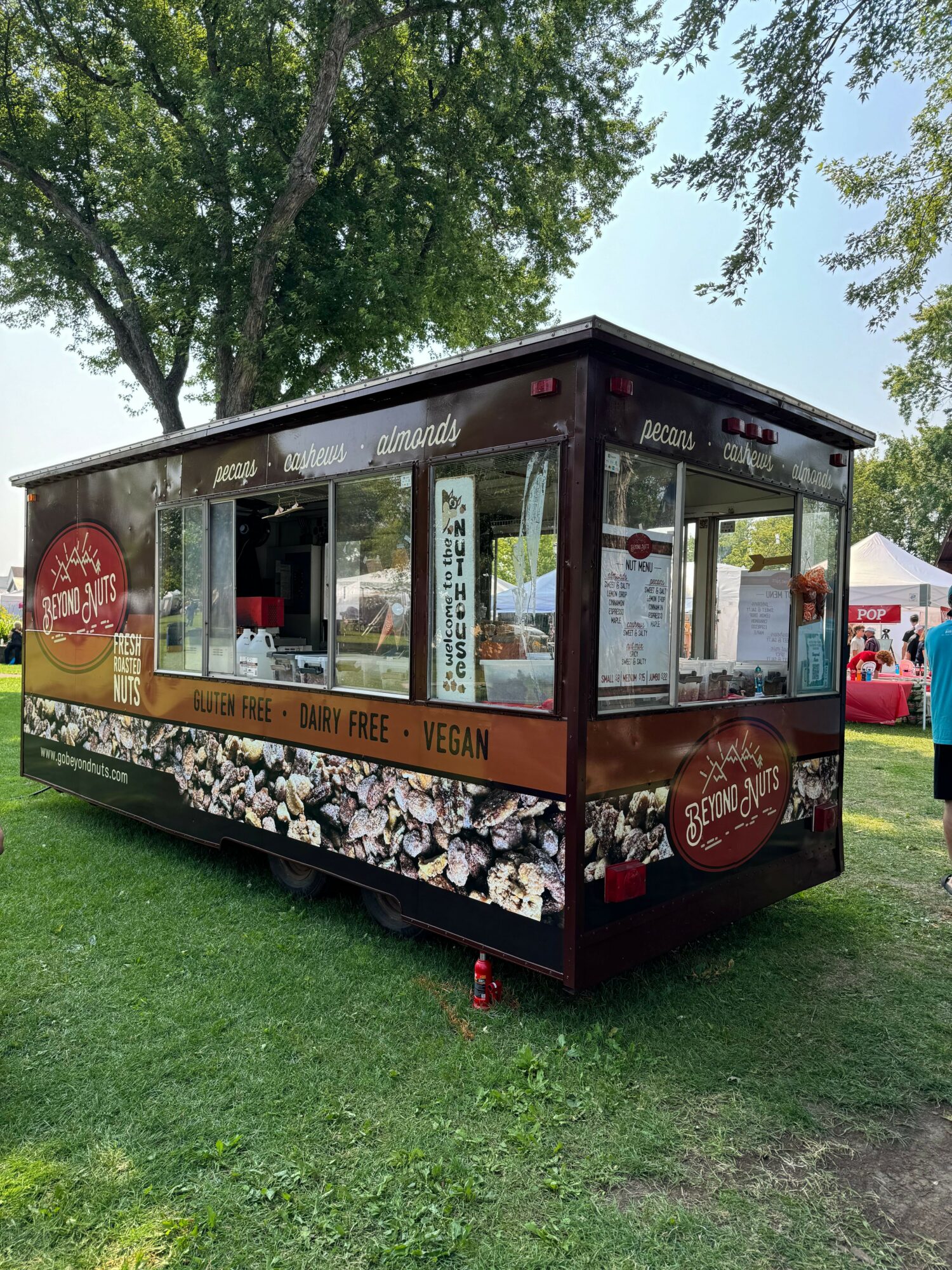 Food truck with large windows, parked on grass with trees and tents in background.