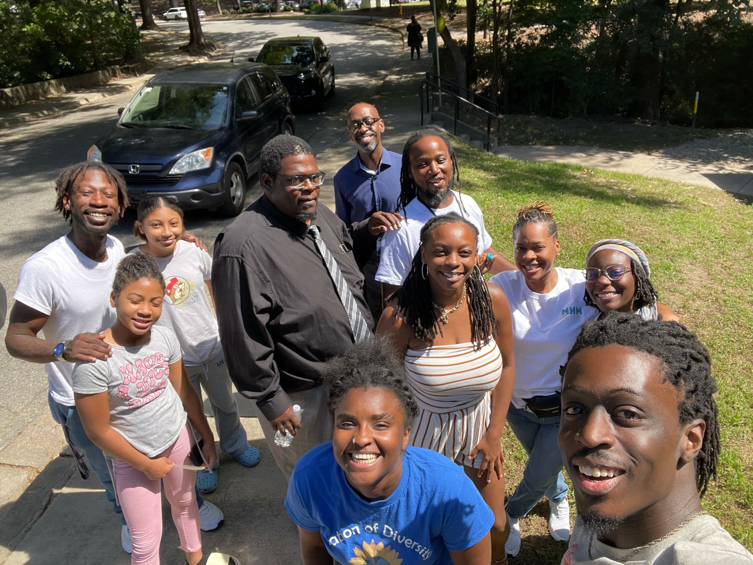 Group of ten people smiling outdoors on a sunny day, standing on grass and pavement, with cars and trees in background.