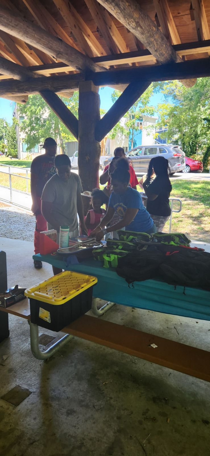 Group of people standing outdoors under a wooden shelter, with trees and parked cars in the background.
