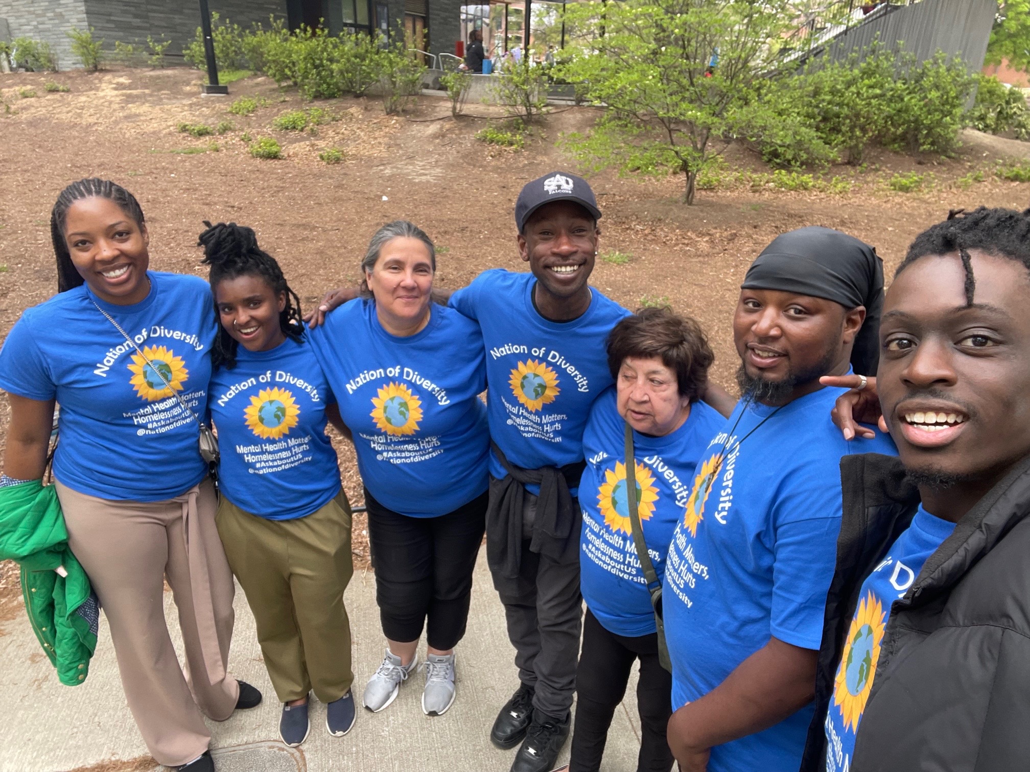 Group of seven people outdoors, wearing matching blue shirts with a yellow sunflower logo, smiling and standing close together.