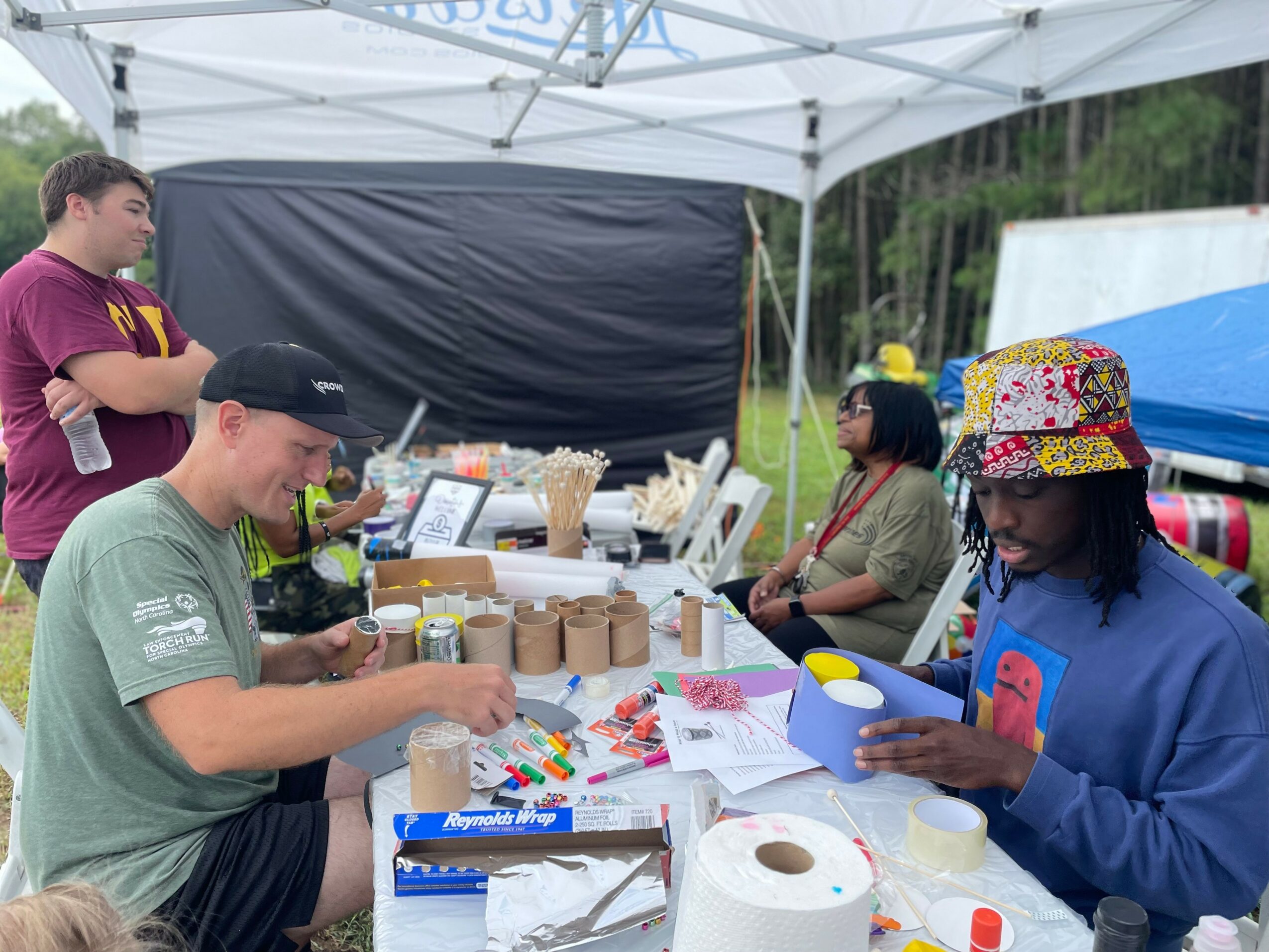 People at an outdoor booth with supplies, some seated and one standing, under a white canopy, with trees in background.