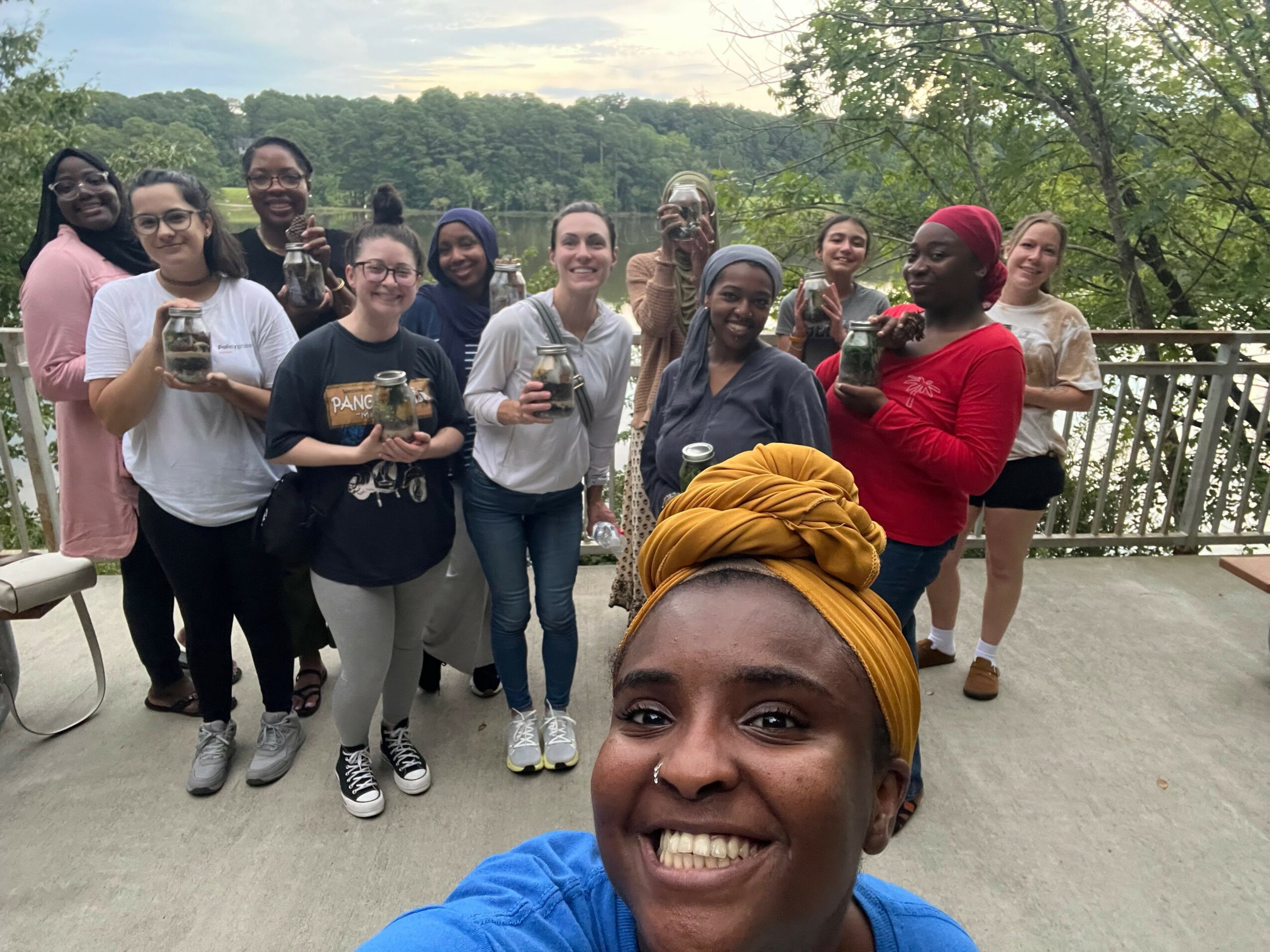Group of people standing outdoors on a balcony, smiling, holding drinks, with trees and sky in background.