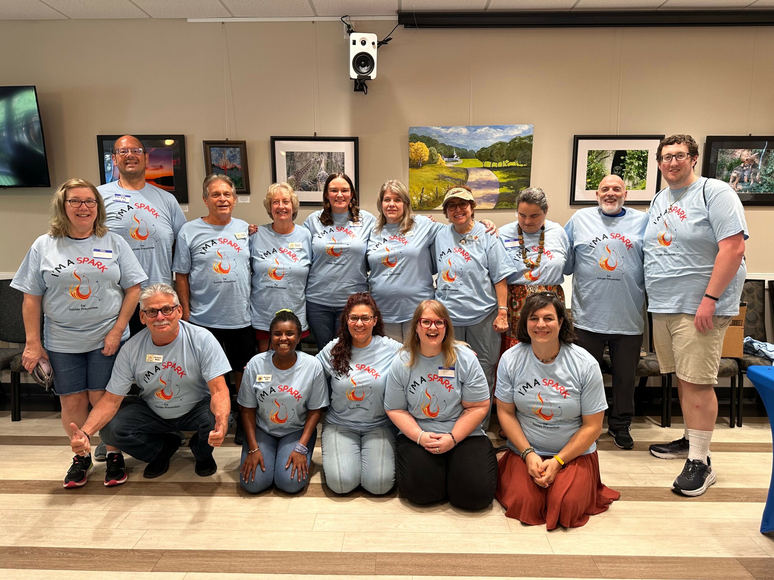 Group of fifteen people in matching light blue shirts, posing indoors with framed pictures on the wall behind them.