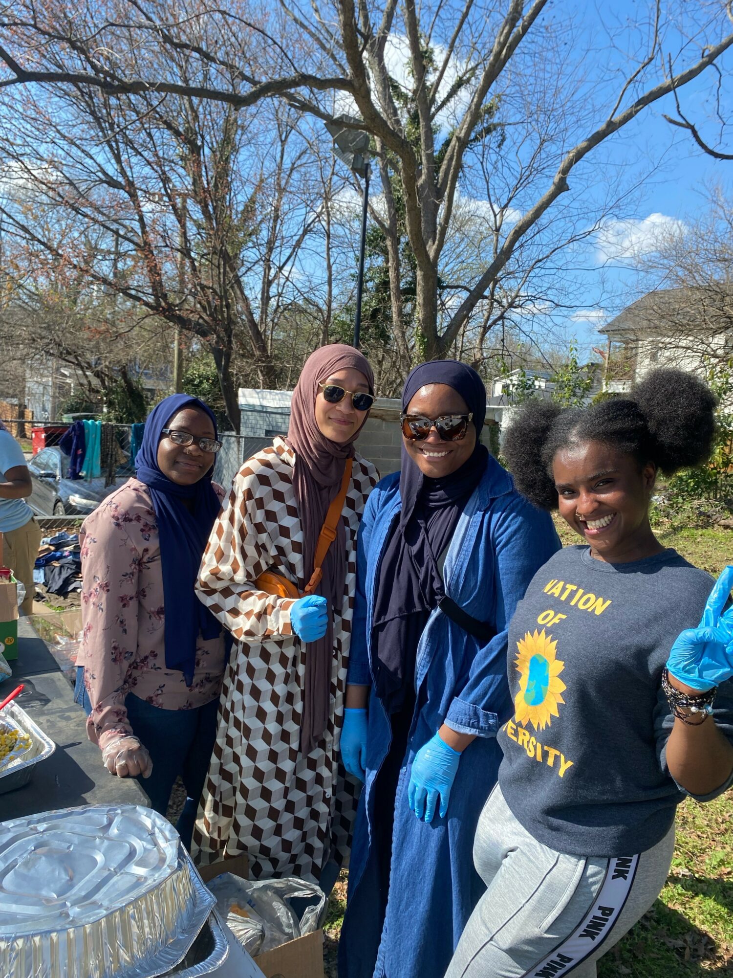 Four women standing outdoors, smiling, wearing sunglasses and gloves, with trees and a house in the background.